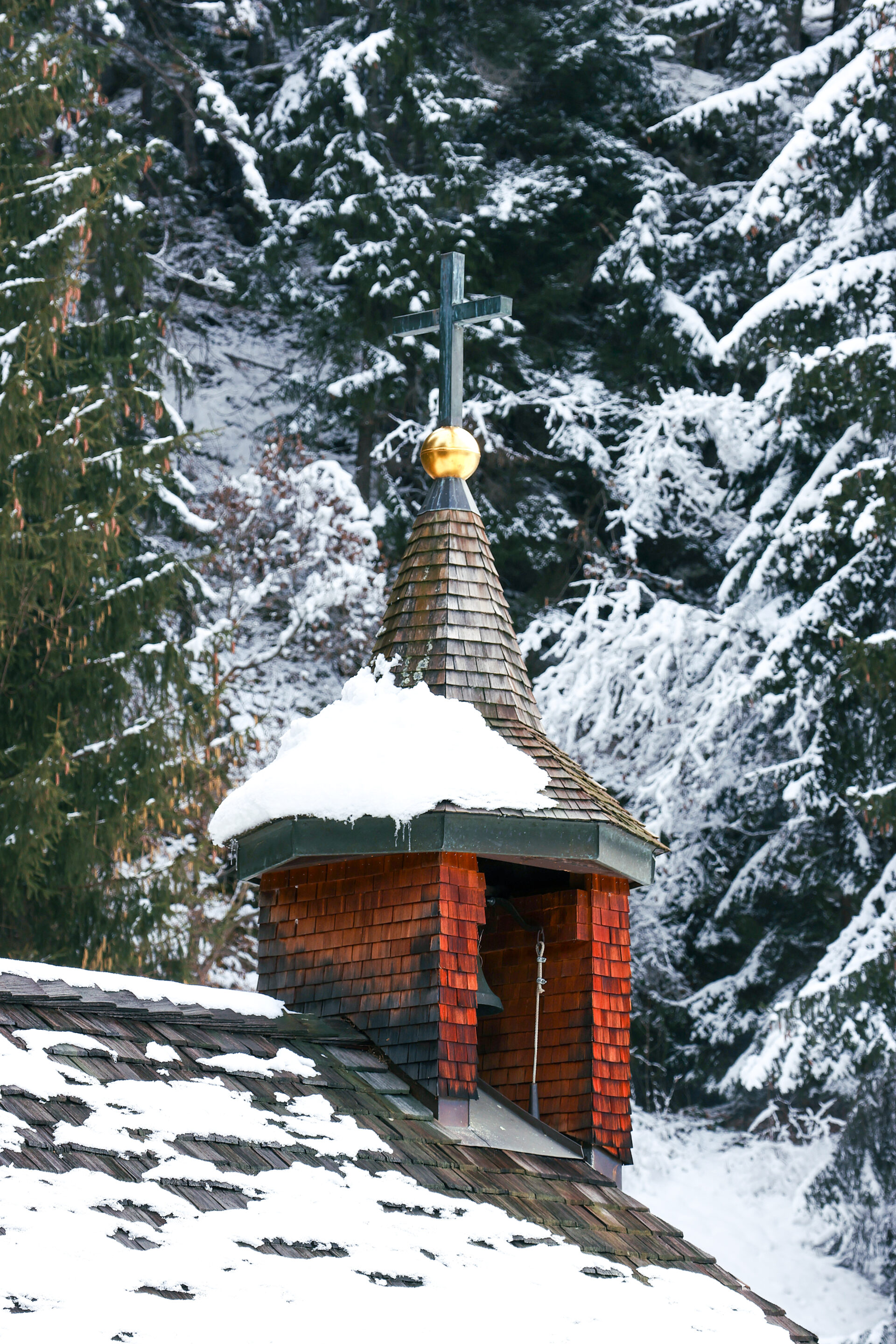 Das Bild zeigt den kleinen Glockenturm einer Kapelle mit hölzernen Schindeln, auf dessen Dach eine dicke Schneeschicht liegt. Ein Kreuz mit goldener Kugel krönt den Turm, während im Hintergrund verschneite Tannen eine friedliche Winterstimmung erzeugen.