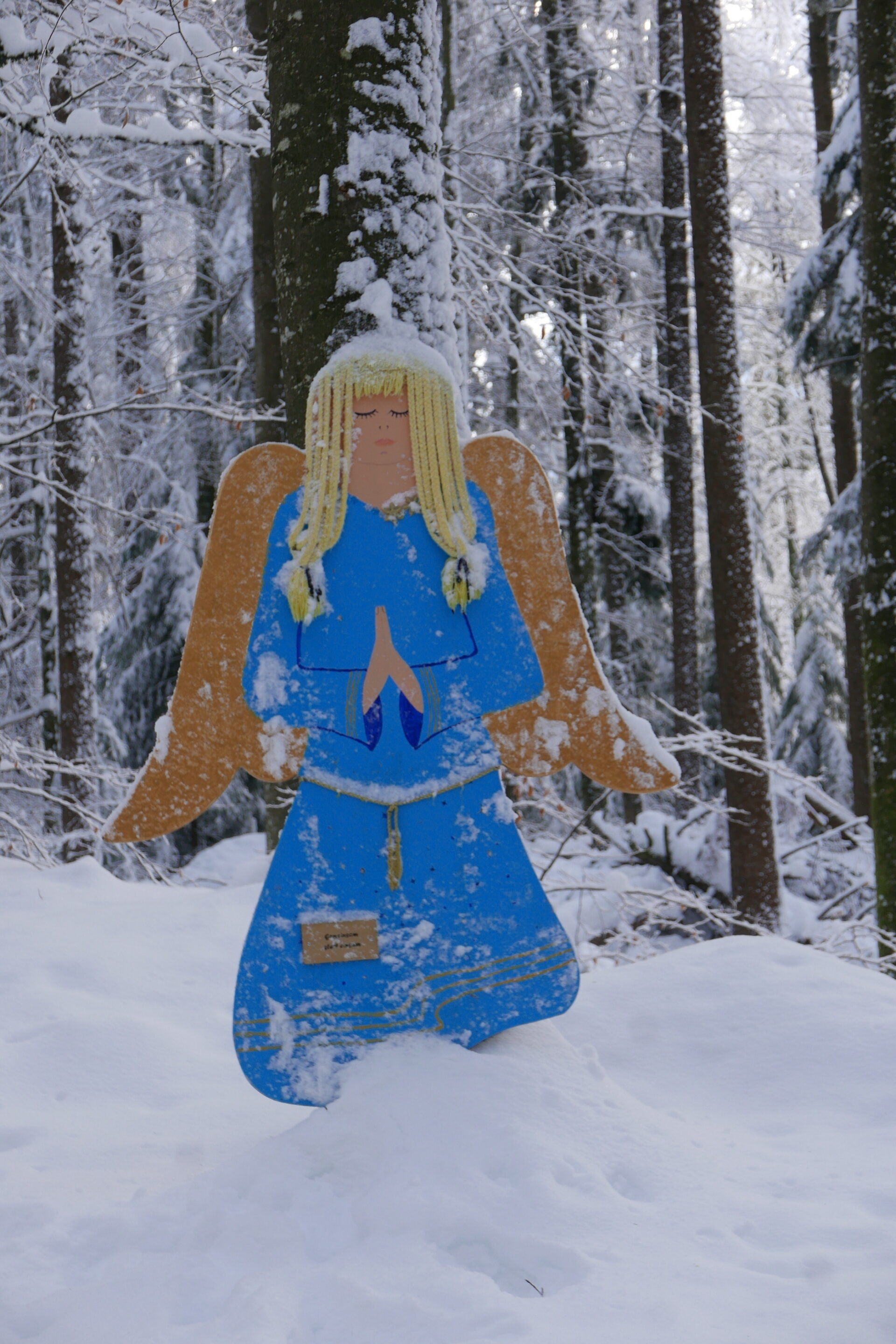 Eine bemalte Holzfigur eines Engels mit blonden Haaren, blauen Gewand und goldenen Flügeln steht im verschneiten Wald. Umgeben von hohen Bäumen und frischem Schnee wirkt die Szene ruhig und winterlich, mit einem Hauch von weihnachtlicher Stimmung.
