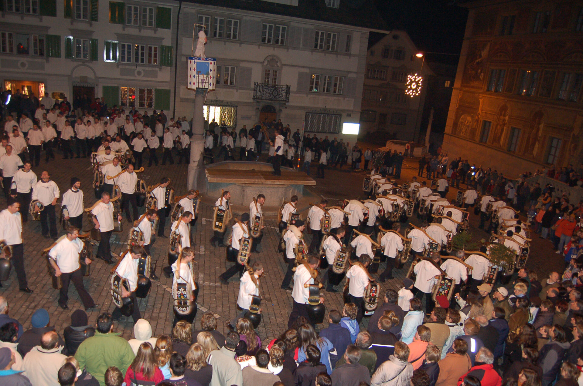 Zuschauer die am frühen Morgen einer Gruppe von Treichler zuschaut die auf dem Hauptplatz um den Dorfbrunnen im Kreis läuft