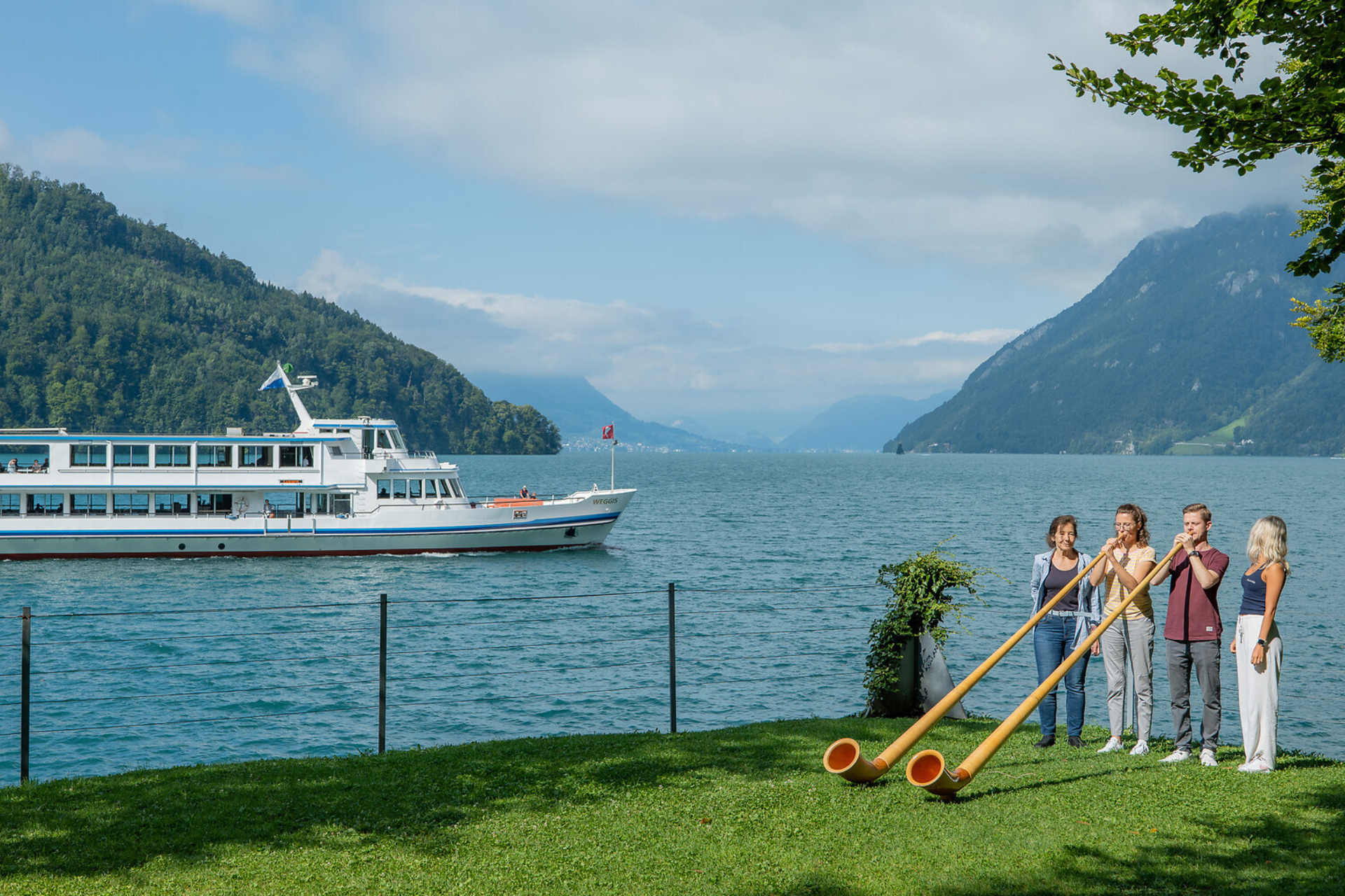 Vier Personen spielen Alphorn am Ufer des Vierwaldstättersees, während ein Passagierschiff auf dem türkisblauen Wasser zwischen bewaldeten Bergen fährt.