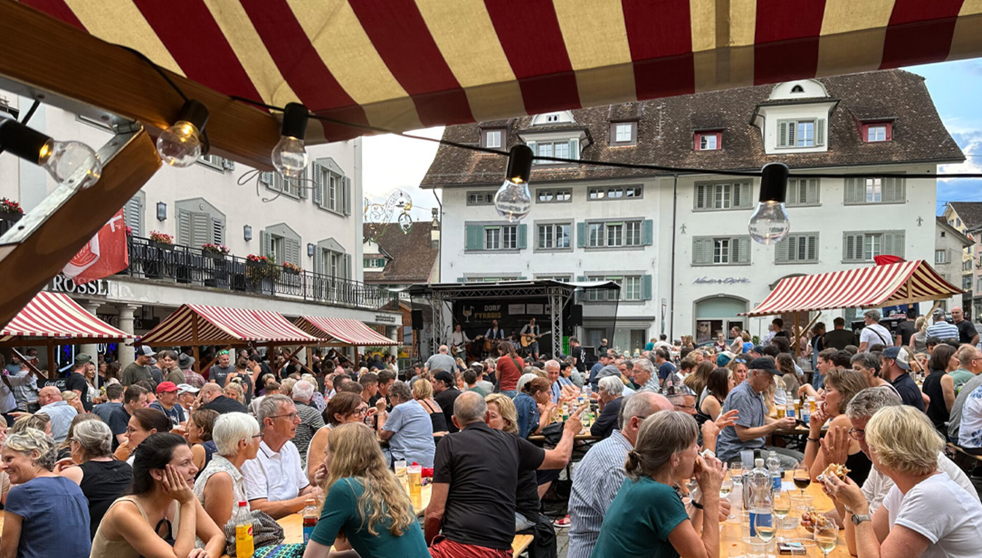 eine grosse Menschenmenge die auf dem Hauptplatz in Schwyz an Tischgarnituren sitzt und miteinander Sprechen wären im Hintergrund eine Band spielt