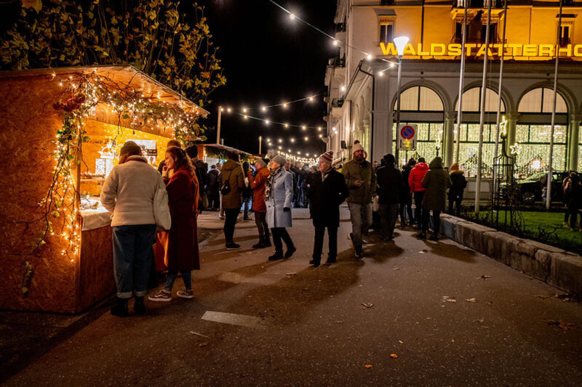 Menschen schlendern gemütlich über einen festlich beleuchteten Weihnachtsmarkt, wobei ein Stand mit Lichtern und Dekorationen die Aufmerksamkeit auf sich zieht. Im Hintergrund leuchtet das "Waldstätter Hof" Hotel in warmem Licht, während Lichterketten die nächtliche Szene stimmungsvoll ergänzen.