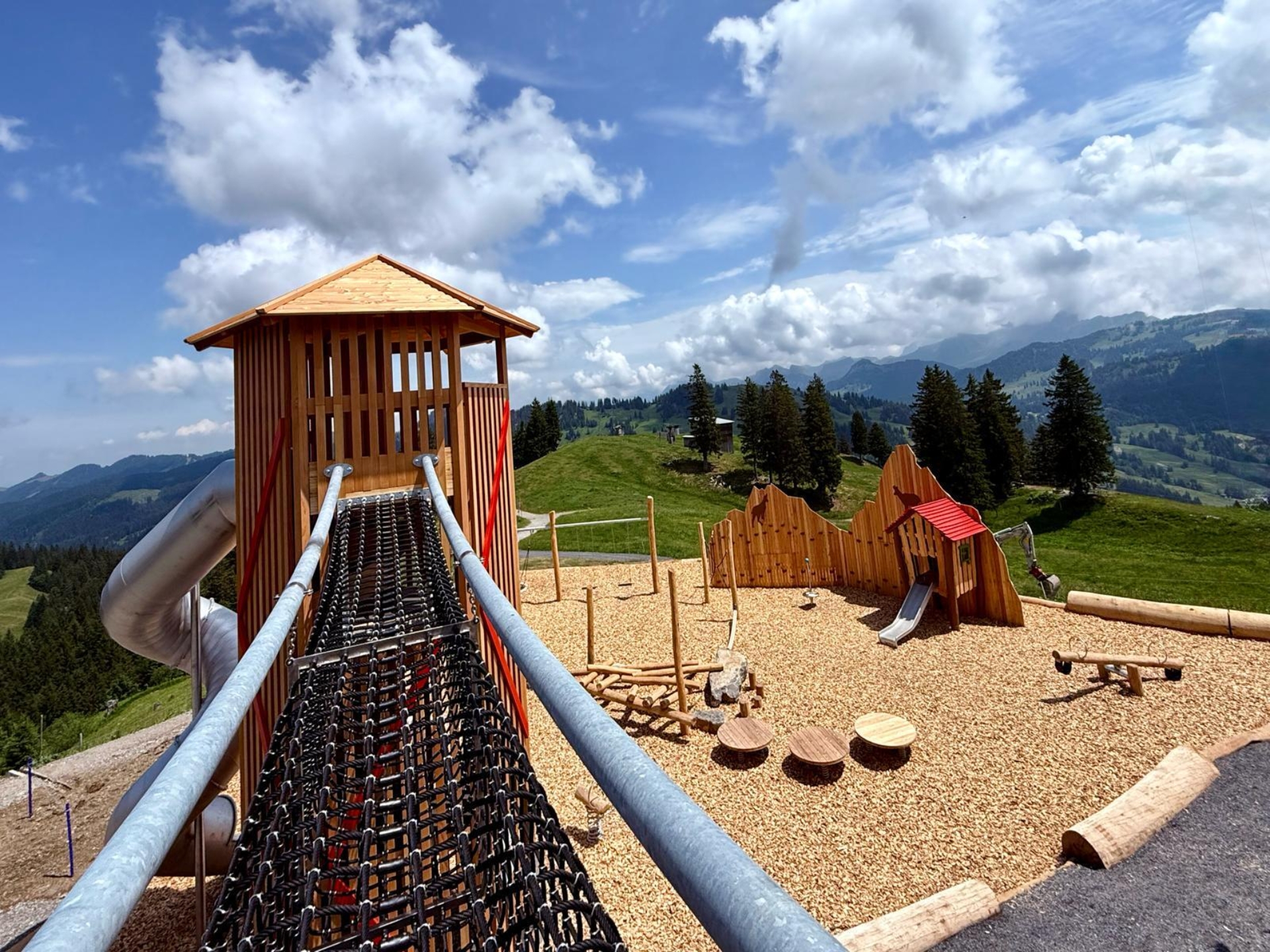 Spielplatz Gämsipark auf der Rotenflue im Sommer. Blick über die Hängebrücke welche zum Holz-Kletterturm führt. Hinter dem Turm stehen Schaukeln und Kletterelemente.