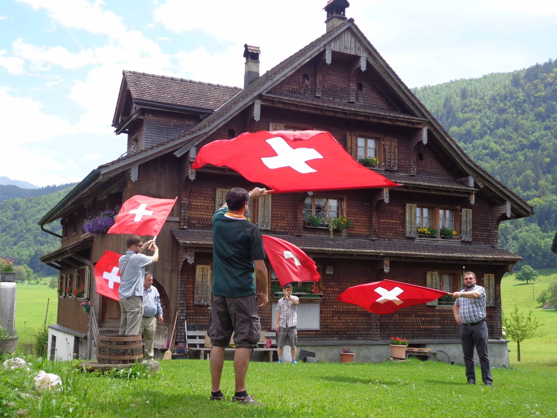 Eine Gruppe von 5 Erwachsenen schwingt grosser Schweizerfahnen vor einen traditionellen Schwyzer Holzhaus. Das Haus steht auf der grünen Wiese. Der Himmel ist blau und leicht bewölkt.