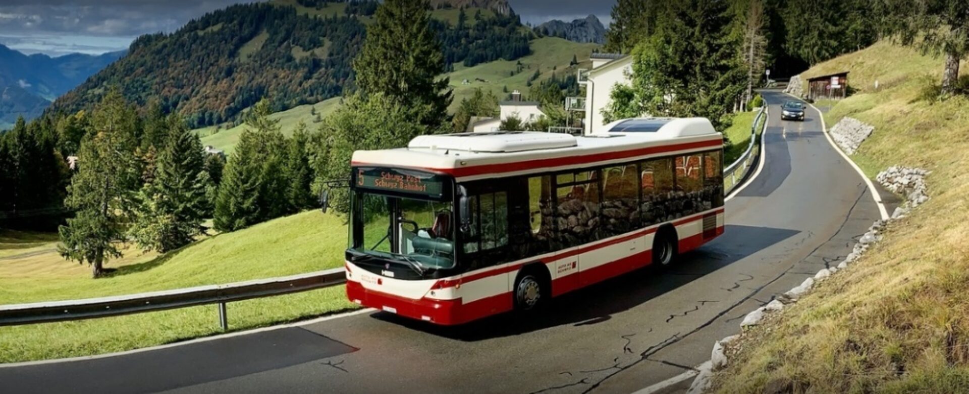 Der rote Bus der Auto AG Schwyz fährt die Ibergeregg-Strasse hinunter. Die Strasse wird von grünen Matten umgeben und mehrere Tannenbäume und ein Haus sind im Hintergrund sichtbar.