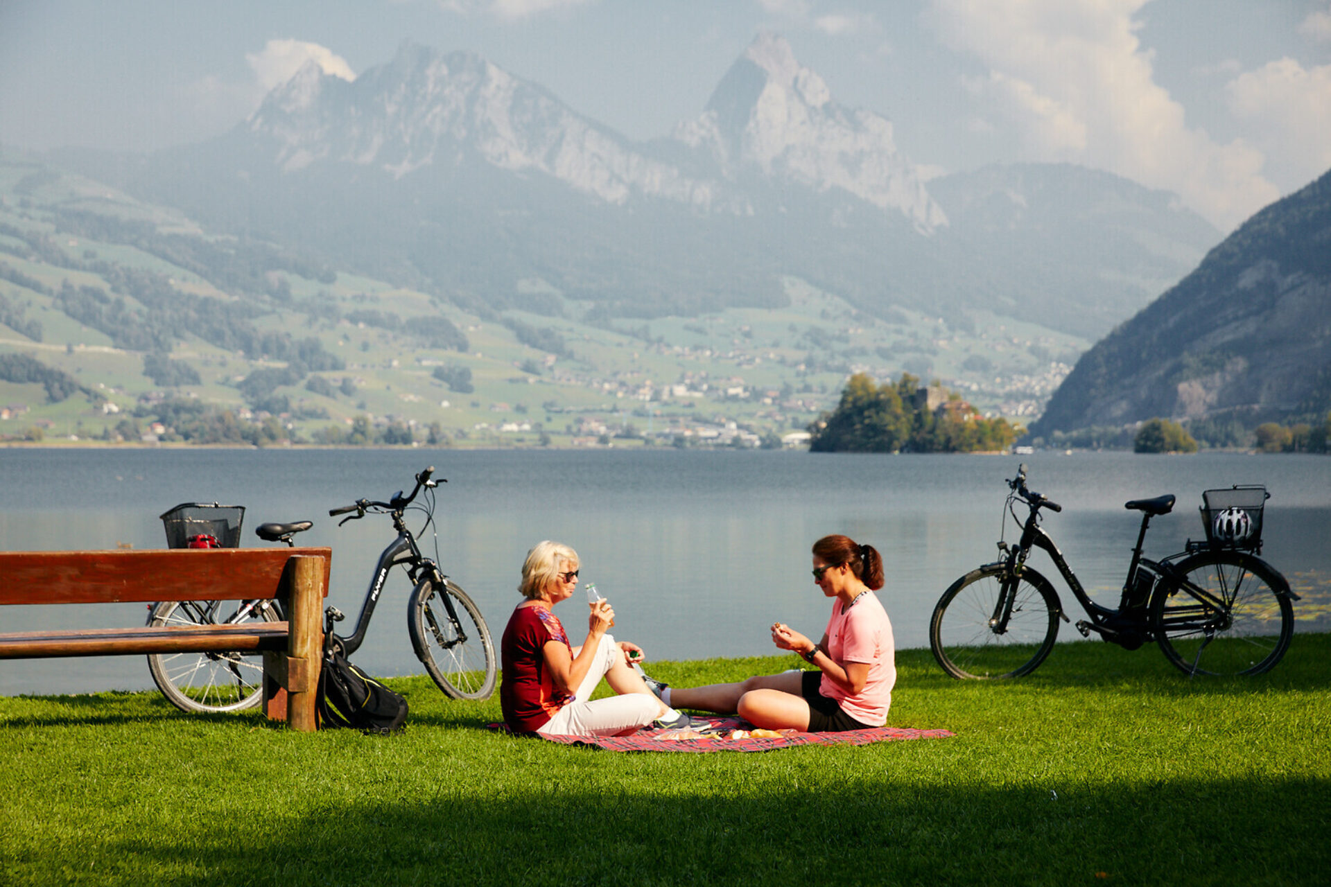 Zwei Damen geniessen ein Picknick am Lauerzersee mit Blick auf die Mythen. Je links und rechts von den Damen steht ein E-Bike.