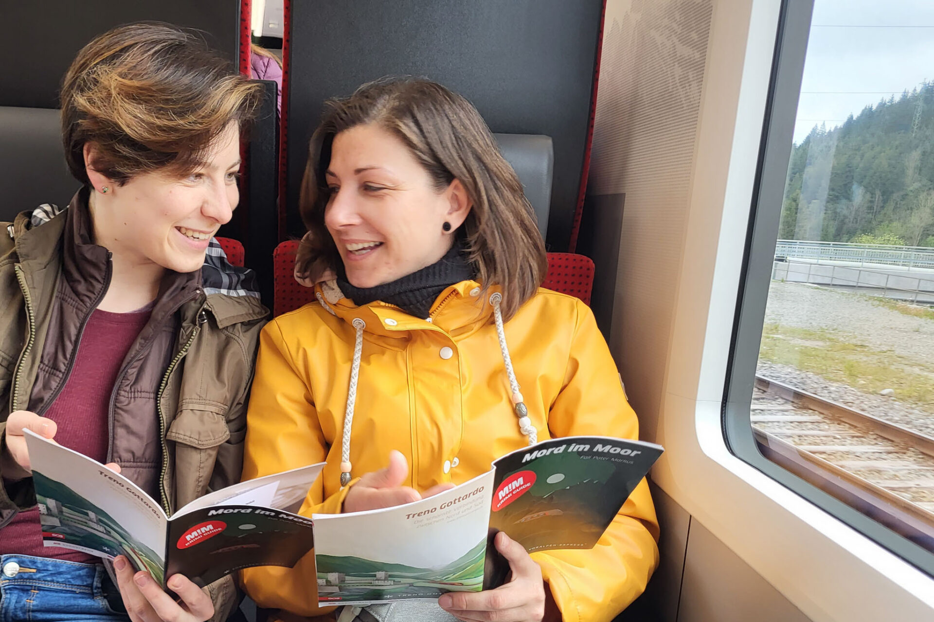 Zwei Damen sitzen im Zug "Voralpen Express" der SOB. Sie sind in Regenjacken gekleidet und lesen zusammen eine Broschüre zum Angebot "Mord im Moor". Durch das Fenster sind Schienen und Wald zu sehen.