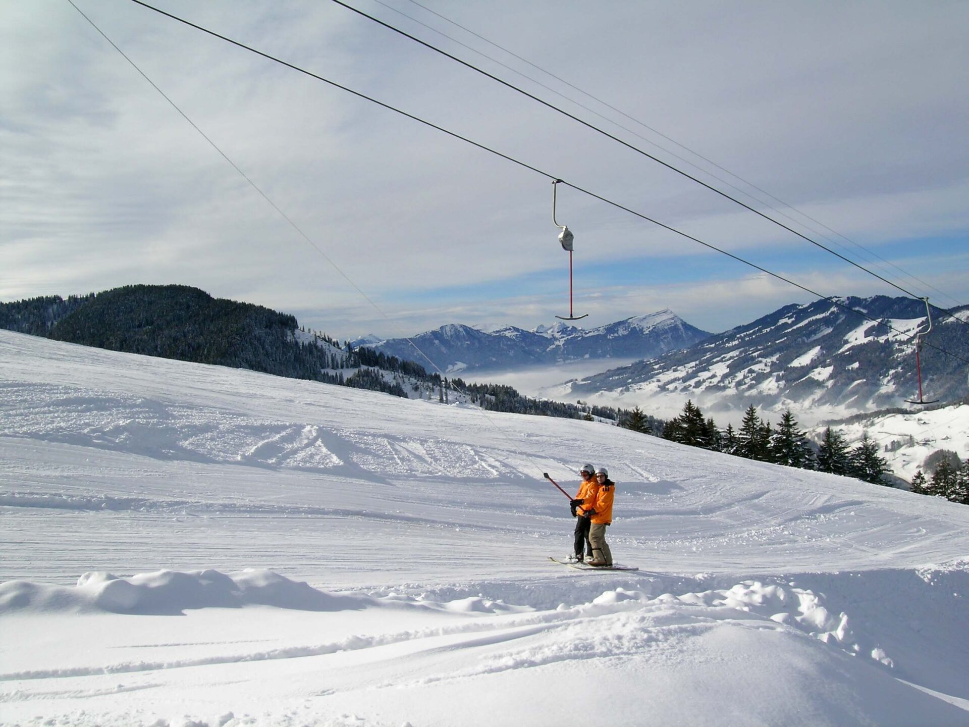 Am Skilift Neusell in Rothenthurm: Eine schön verschneite Winterlandschaft. Die Zugseile des Skilifts sind sichtbar. Ein Bügel bringt zwei Snowboarder hoch. Beide tragen orange Jacken und Helm.