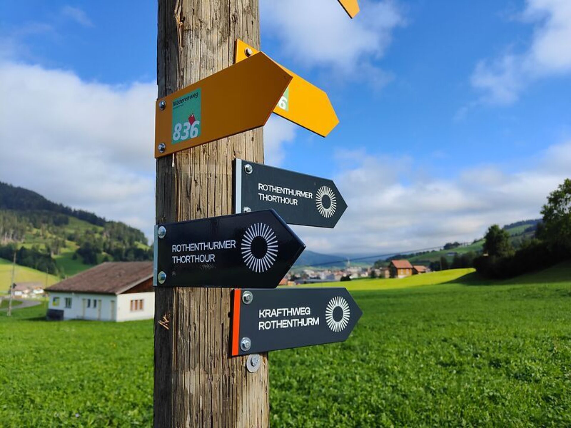 Signalisation Wanderwege Rothenthurm: Gelbe und schwarze Wegweiser an einem Holzmasten. Der Wegweiser steht in einer grünen Wiese. Im Hintergrund sind einige Häuser von Rothenthurm sichtbar. Der Himmel ist blau und mit weissen Wolken durchzogen.