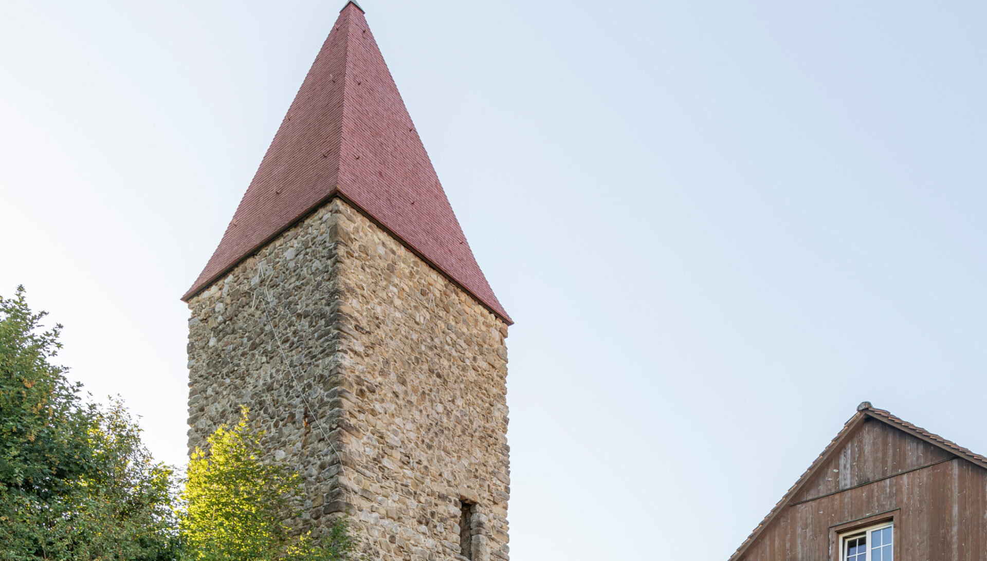 Vom historischen Letziturm in Rothenthurm ist der ober Teil und das rote Dach sichtbar. Der Turm ist aus Stein und an der Fassade sind die einzelnen Steine sichtbar. Ein einziges Fensterloch ist in der Mitte unten ersichtlich. Neben dem Turm sieht man den Dachstock eines Holzhauses. Der Himmel ist hellblau.