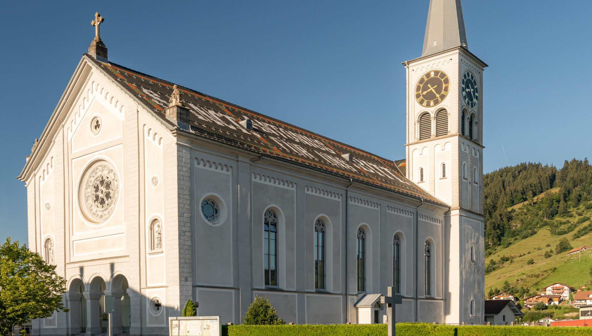 Pfarrkirche St. Antonius Rothenthurm. Eine grosse Pfarrkirche aus weissem Sandstein. Die Vorderfront ist verziert und hat ein grosses Rundes Fenster. Rechts ragt der Kirchturm mit einem spitzen Dach hoch. Auf dem Dach der Kirche ist eine grosse Inschrift leicht sichtbar. Der Himmel ist strahlend blau.