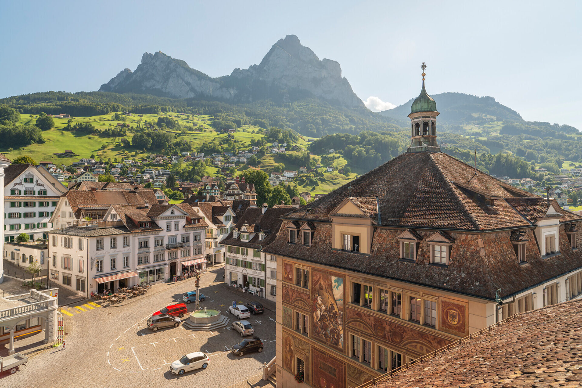 Blick auf den Hauptplatz Schwyz mit dem Grossen Rathaus und dem Dorfbrunnen. Hinter dem Dorf geht es über grüne Wiesen hoch bis zum Grossen und Kleinen Mythen.