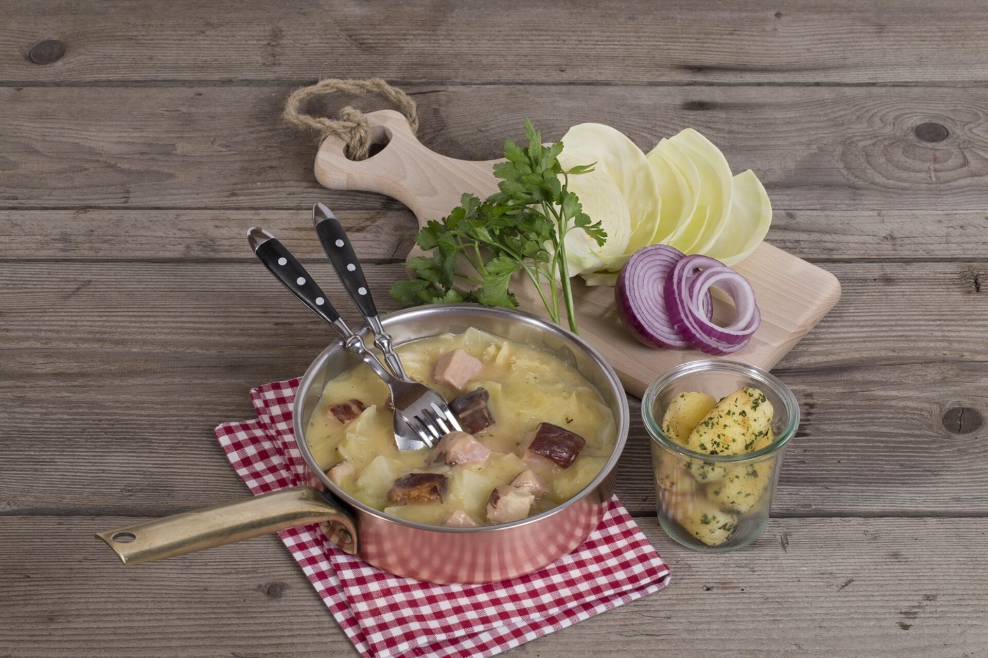 Eine Pfanne voll Hafenchabis schmackhaft angerichtet auf rotweiss Karierten Serviette. Daneben dekorativ Kabisblätter, Zwiebeln und Peterli sowie eine Schüssel Kartoffeln.