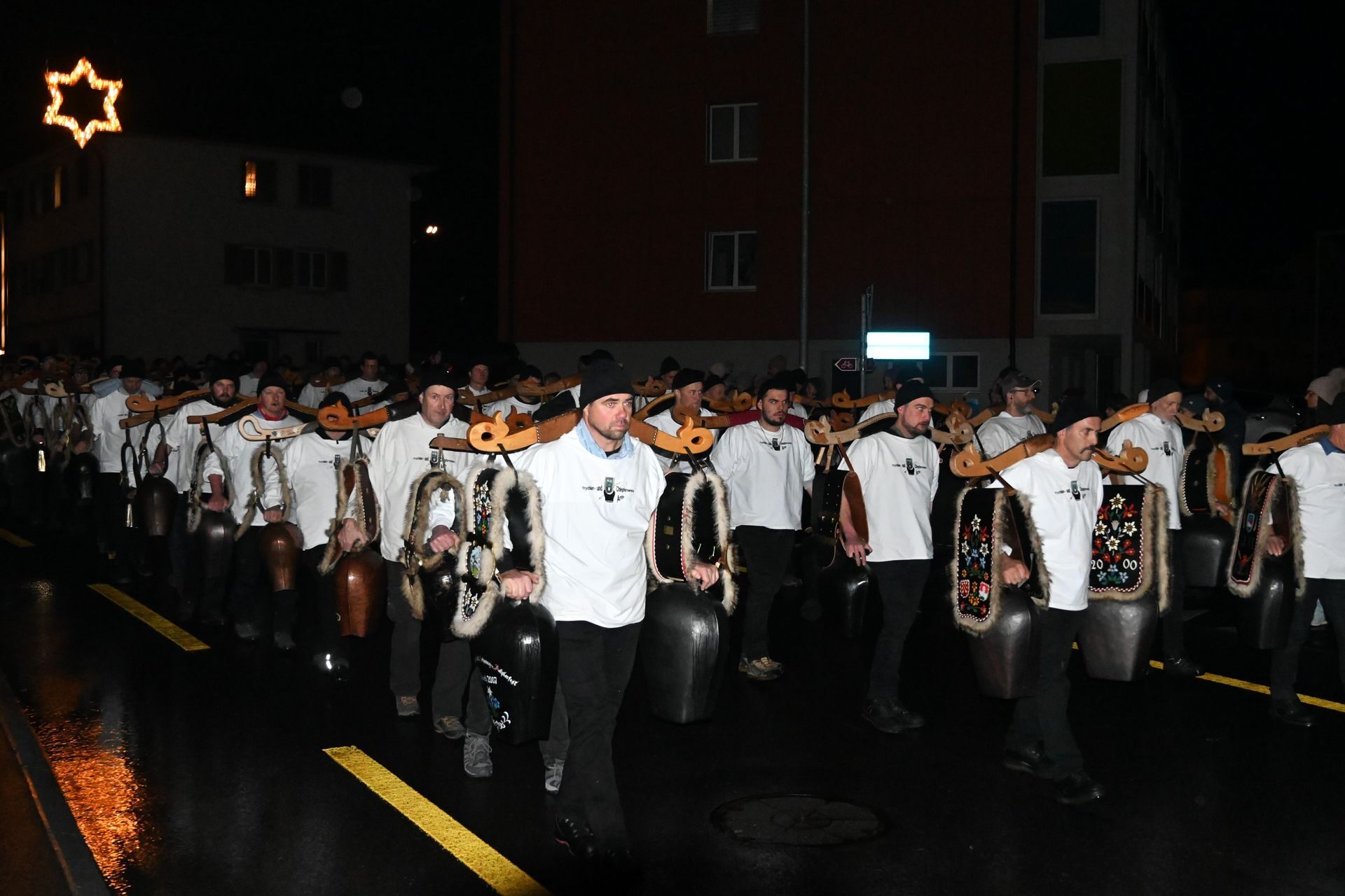 Viele Männer mit weissen Hemden, schwarzer Kappe und einem Joch rechts und links davon baumelt eine Kuhglocke.