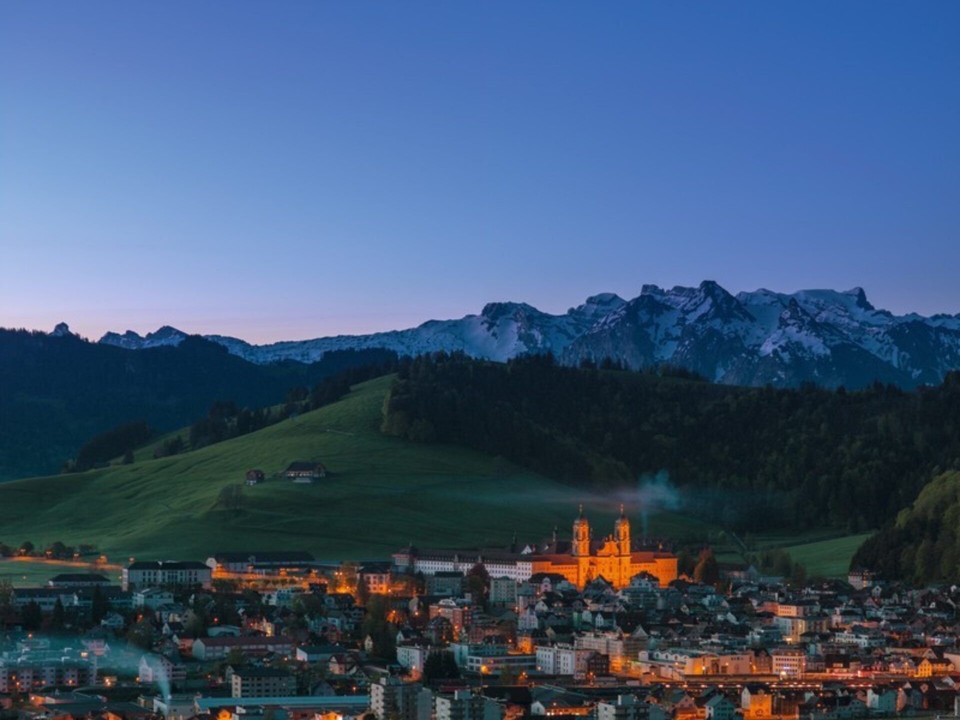 Ortsbild über Einsiedeln mit beleuchtete Kloster und im Hintergrund Schneeberge.