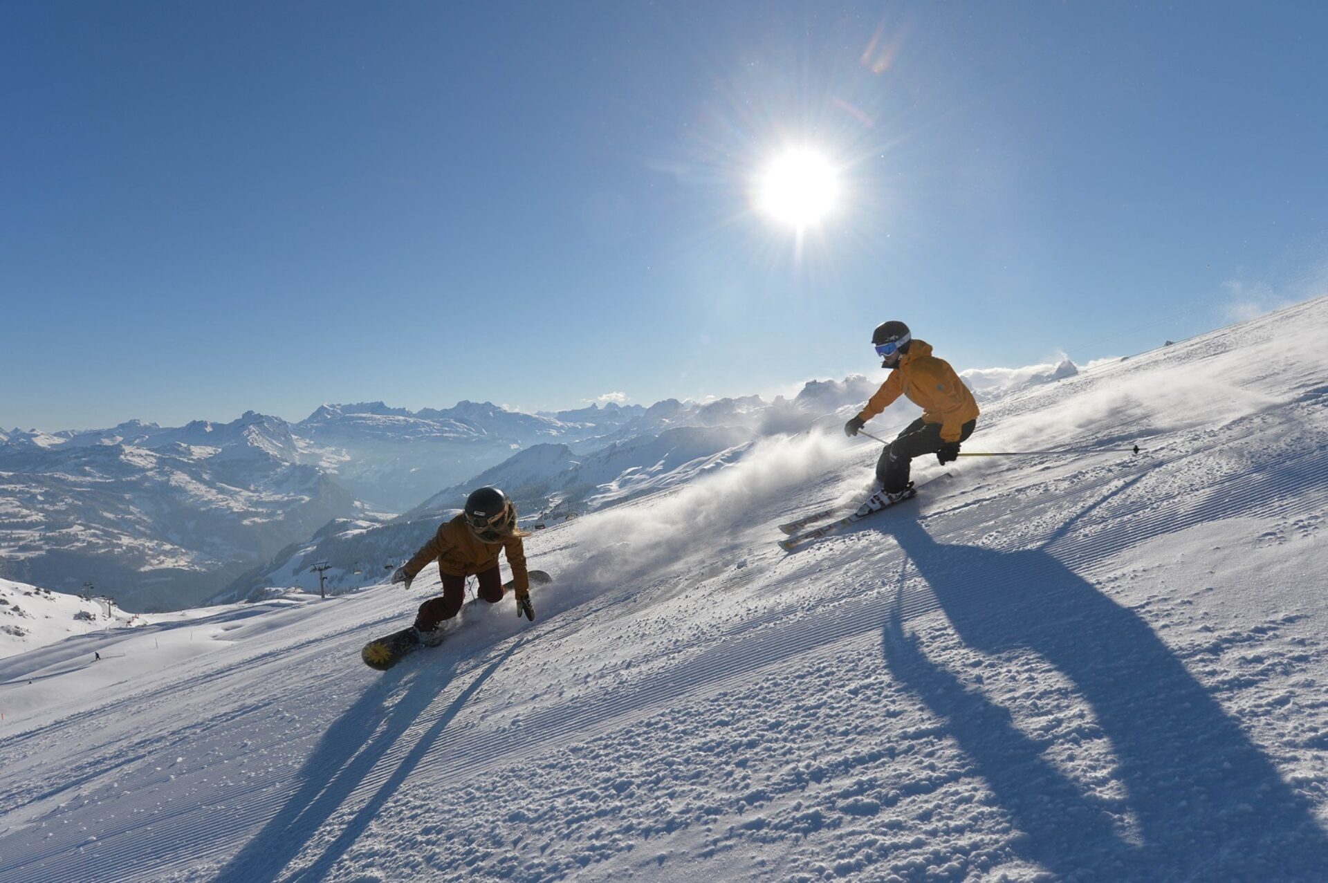 Ein Snowboarder und ein Skifahrer fahren bei Top Verhältnissen die Piste runter. Im Hintergrund die Sonne und tolles Bergpanorama.