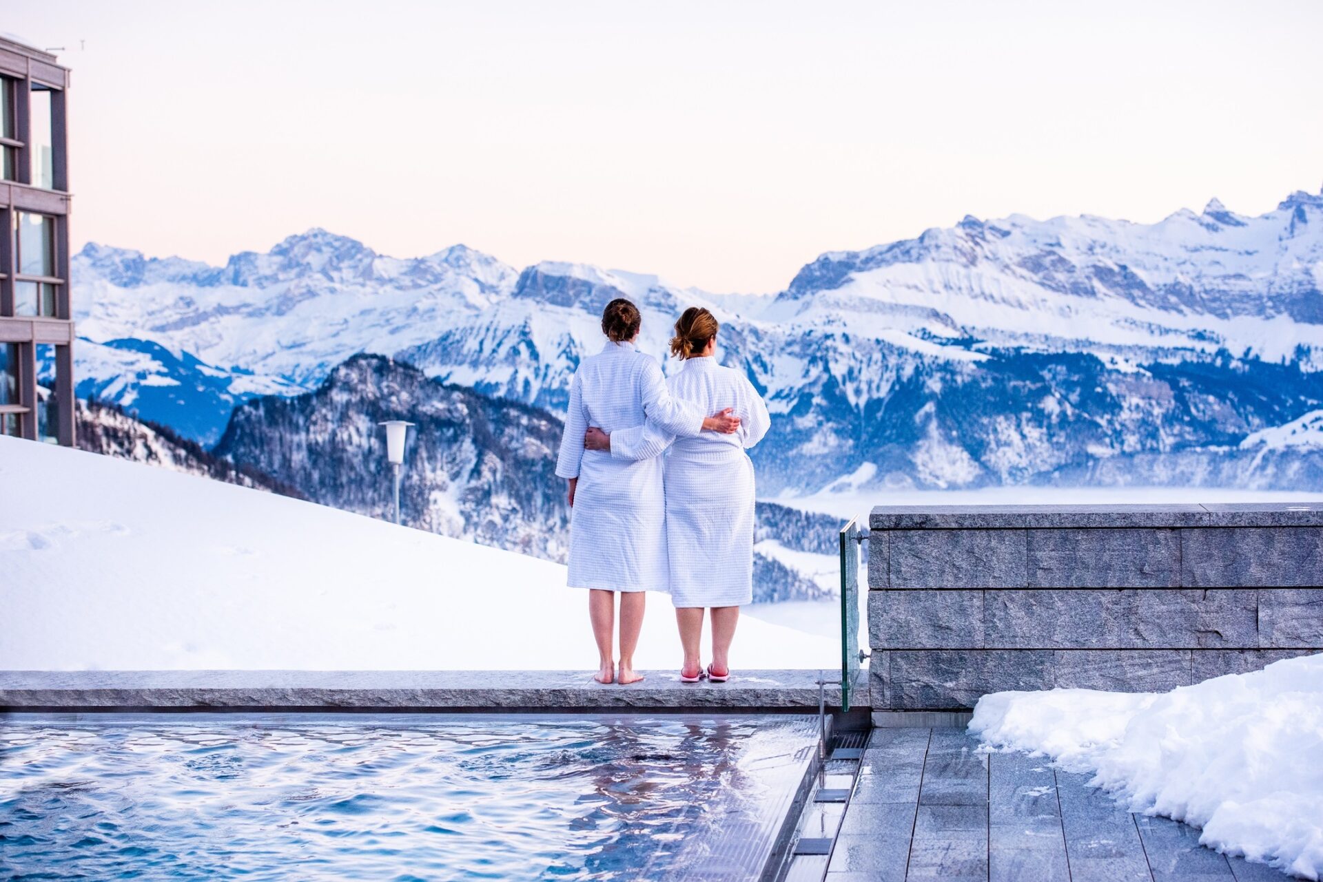 Zwei Frauen in weissen Bademäntel stehen am Poolrand mit Blick zum Berg Panorama.