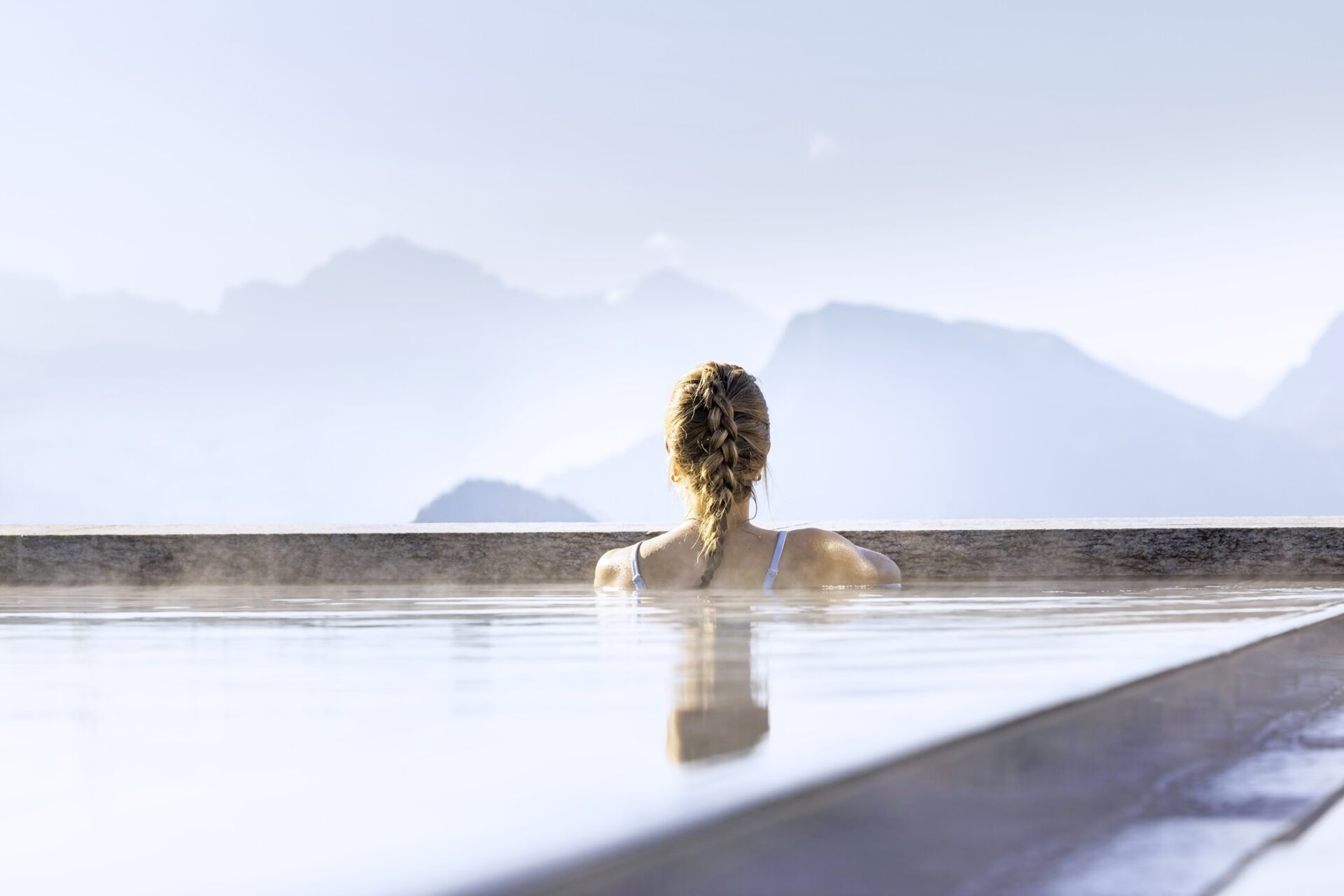 Frau von hinten mit geflüchteten Haaren im Outdoor Pool mit Bergpanoramablick.