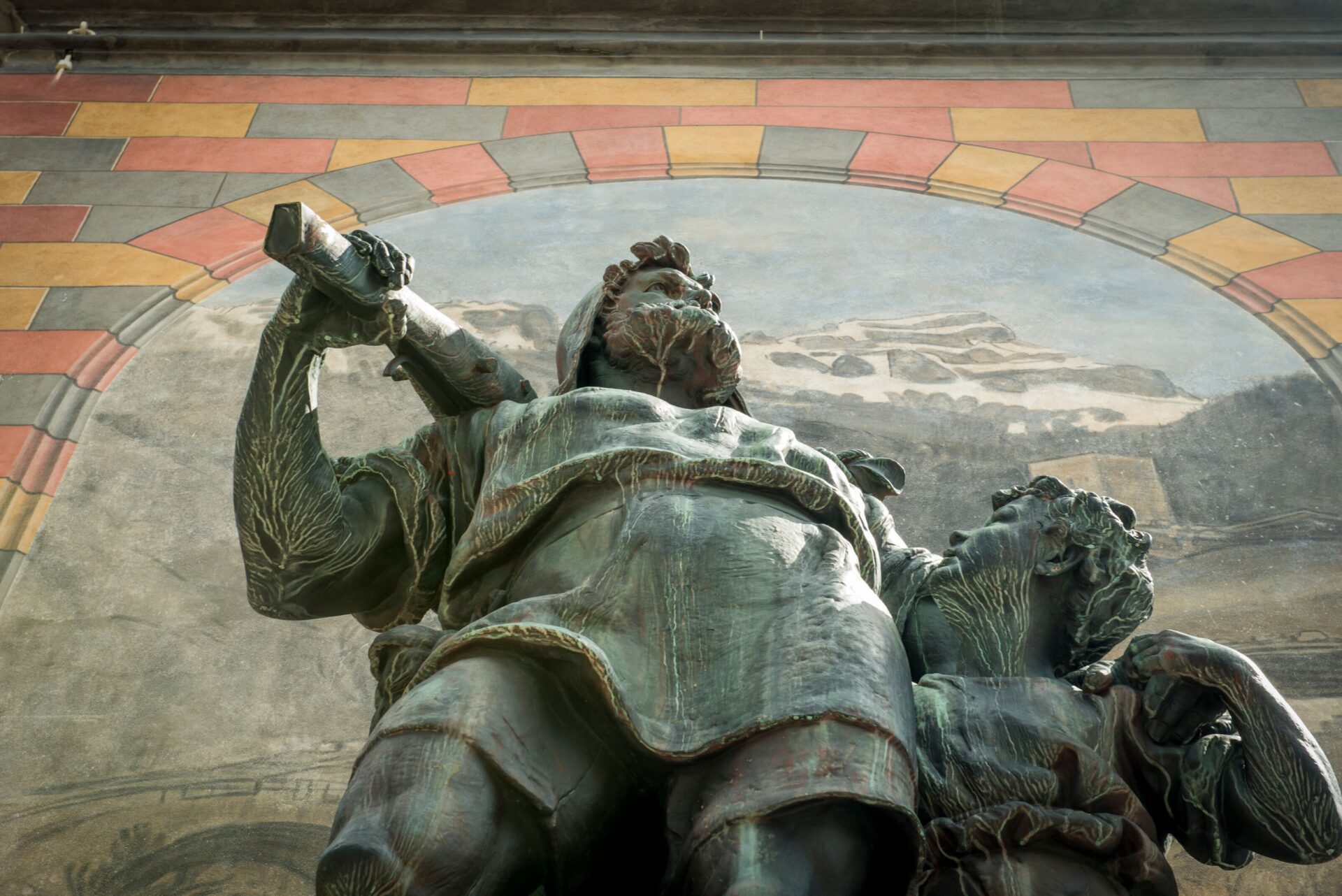 Nahaufnahme einer imposanten Bronze-Statue, die einen Krieger mit entschlossenem Blick und ein Kind an seiner Seite zeigt, aus einer tiefen Perspektive betrachtet. Im Hintergrund ist ein Wandgemälde zu erkennen, das die Szene in einen historischen Kontext einbettet.