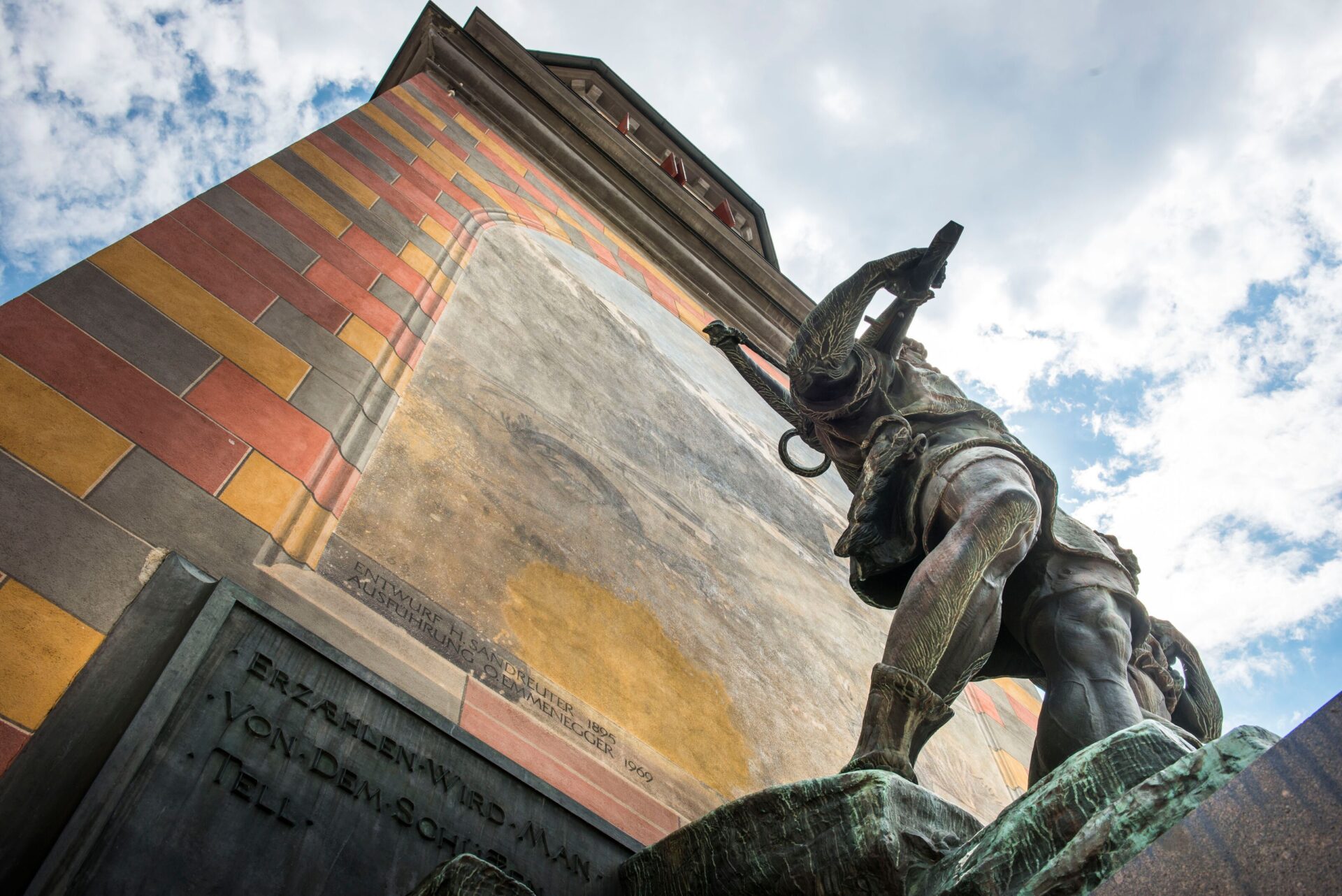 Die Statue von Wilheml Tell wird aus einem dramatischen, tiefen Winkel vor dem bunt gemusterten Turm abgebildet, während der Himmel mit Wolken im Hintergrund den Eindruck von Stärke und Entschlossenheit verstärkt. Diese Perspektive hebt die heroische Pose und die Größe der Figur hervor.