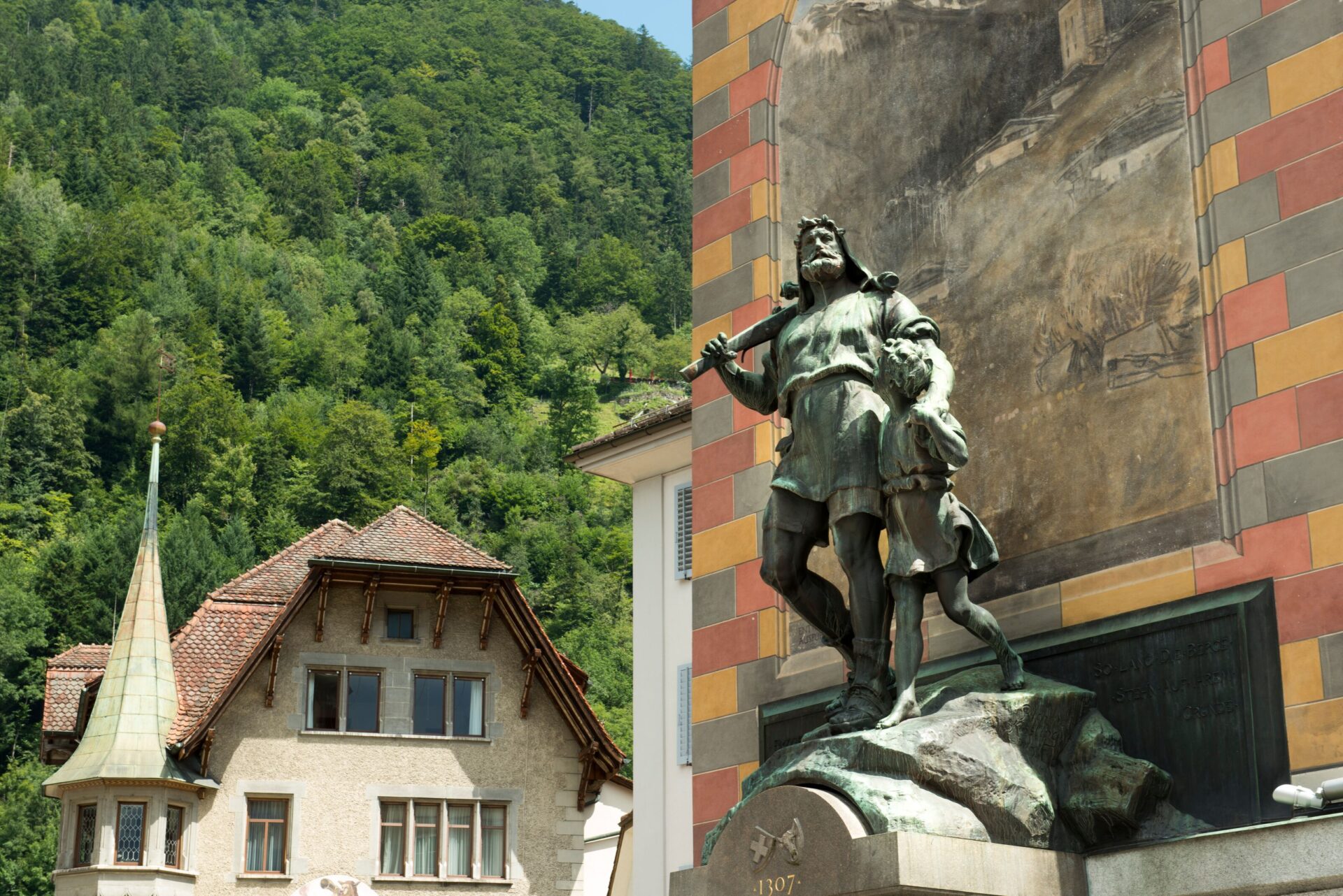 Die Statue von Wilhelm Tell und seinem Sohn steht vor einer bemalten Wand, während im Hintergrund ein Gebäude mit traditioneller Architektur und üppigem Grün zu sehen ist. Das Bild verbindet historische Symbolik mit einer ländlichen Umgebung.