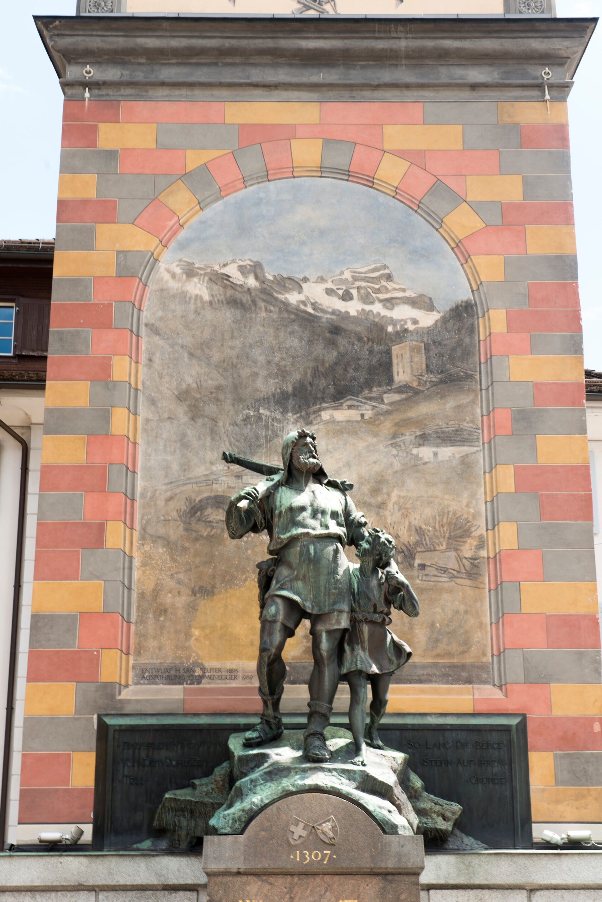 Eine bronzene Statue zeigt einen historischen Krieger mit einem Kind, vermutlich als symbolische Darstellung von Mut und Schutz, vor einer bunt gemusterten Wand mit einem Gemälde von Bergen. Das Kunstwerk kombiniert eindrucksvoll Skulptur und Malerei, um eine patriotische Stimmung zu erzeugen.