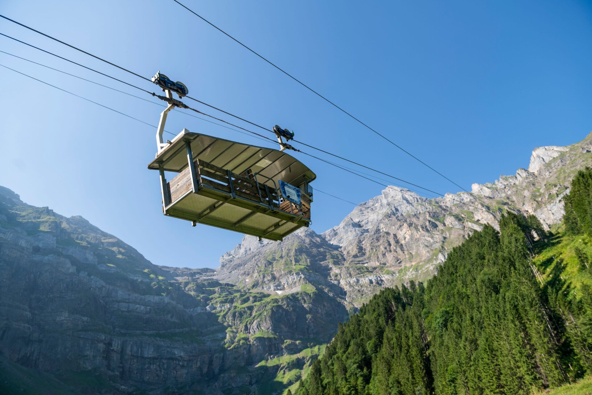 Eine offene Seilbahngondel schwebt hoch über einem grünen Wald, mit steilen Berggipfeln und klarem, blauem Himmel im Hintergrund. Die Szene vermittelt ein Gefühl von Freiheit und Abenteuer inmitten der beeindruckenden Alpenlandschaft.