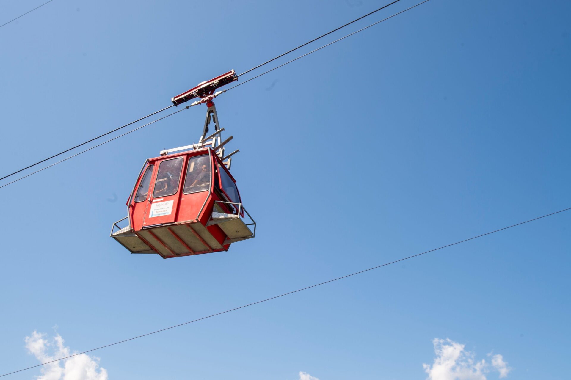 Eine rote Seilbahnkabine schwebt vor strahlend blauem Himmel hoch über dem Boden und vermittelt ein Gefühl von Freiheit und alpiner Leichtigkeit. In der Kabine sind Menschen zu sehen, die die Aussicht genießen und sich auf eine Fahrt durch die Berglandschaft freuen.