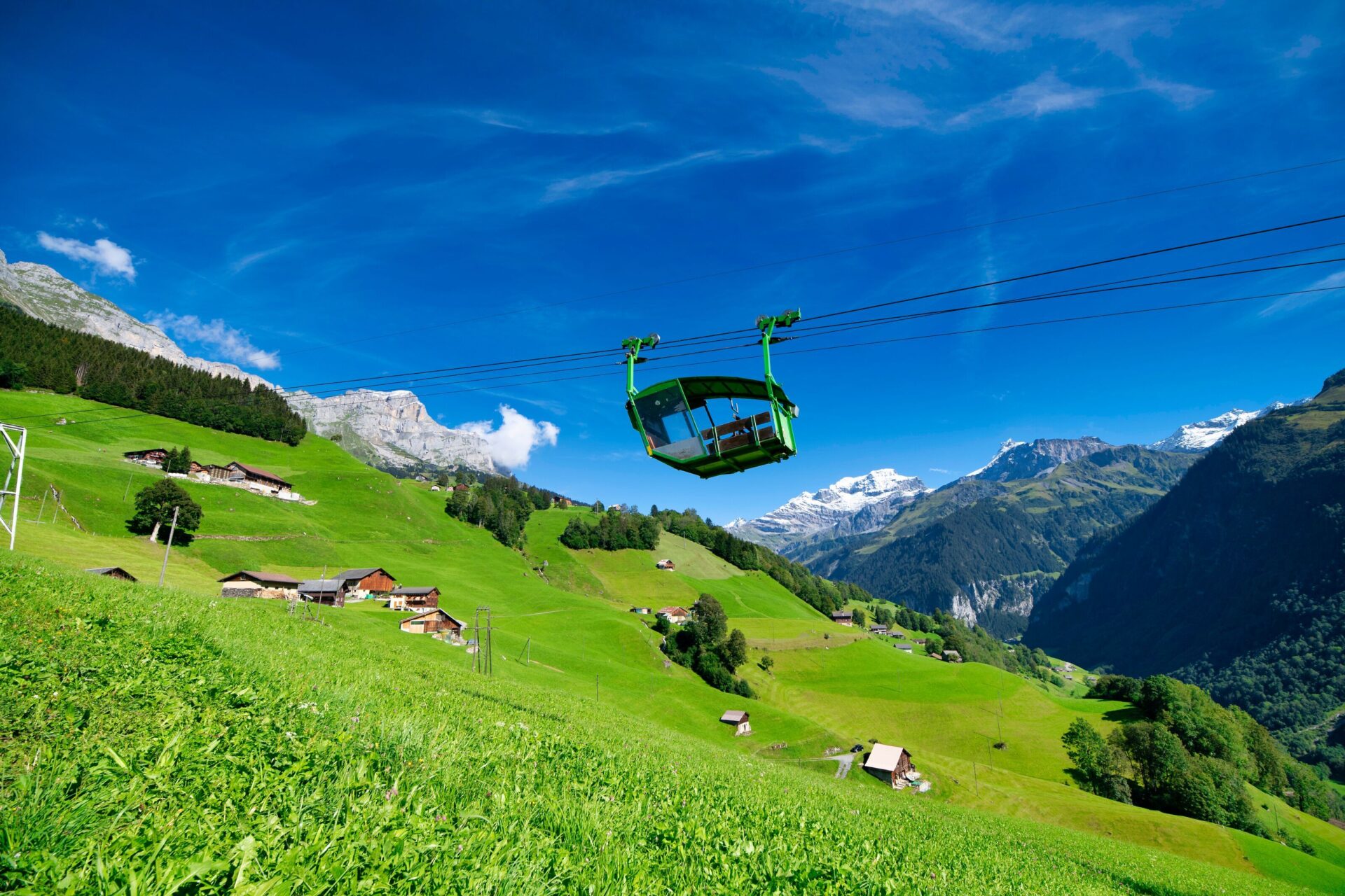 Eine grüne Seilbahngondel schwebt über saftige, grüne Wiesen, mit verstreuten Berghütten und imposanten Gipfeln im Hintergrund unter einem strahlend blauen Himmel. Das Bild vermittelt eine fröhliche und entspannte Atmosphäre inmitten einer malerischen Alpenlandschaft.