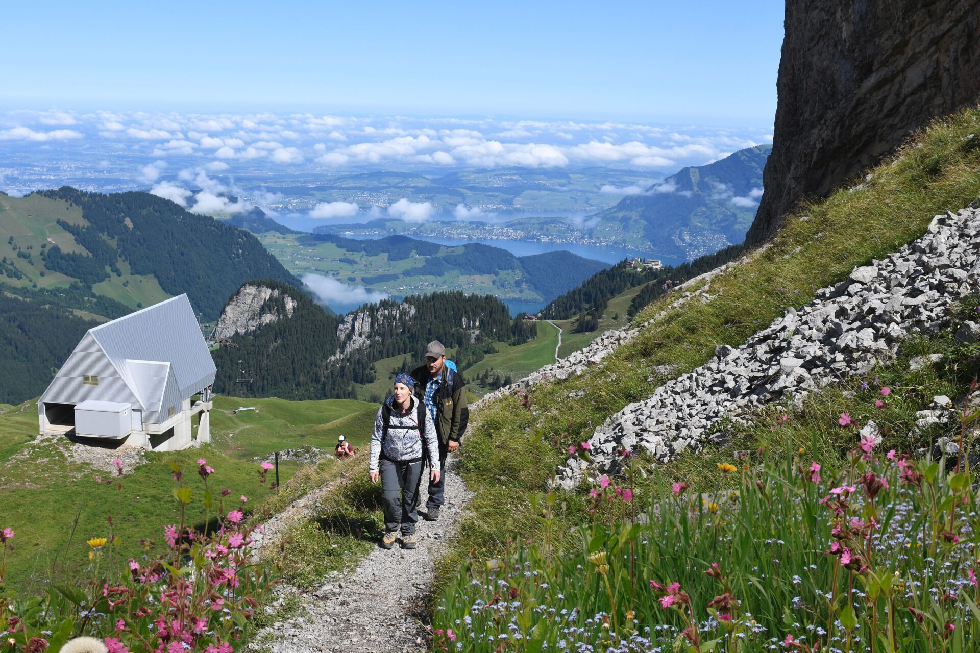 Zwei Wanderer gehen auf einem schmalen Pfad durch eine blühende Alpwiese, während sich im Hintergrund eine moderne Berghütte und eine atemberaubende Berglandschaft erstrecken. Die Szene vermittelt Ruhe, Naturverbundenheit und die Freude am alpinen Wandern.