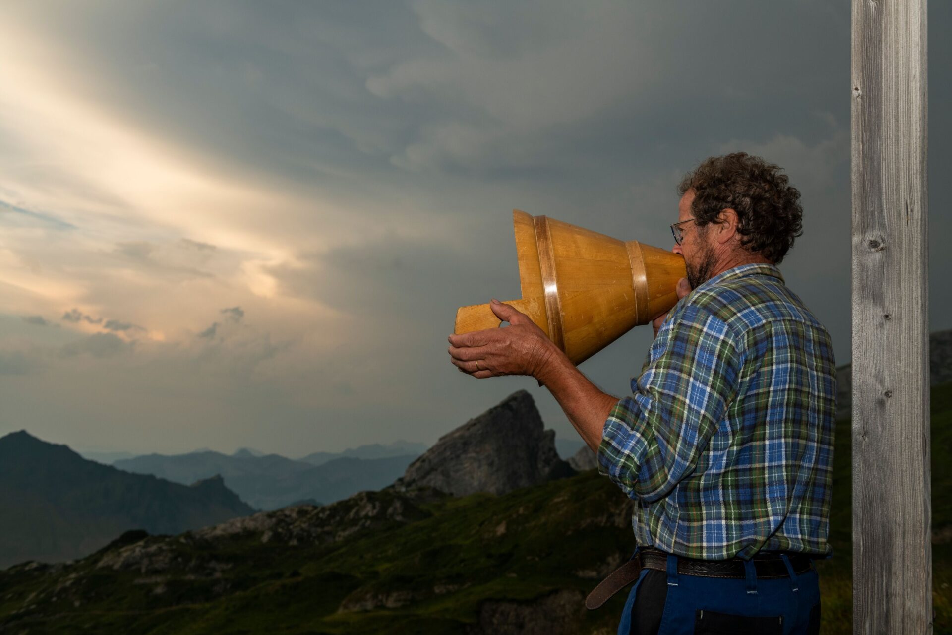 Ein Senn ruft mit einem traditionellen Kuhhorn über die Alpenlandschaft, während die Abenddämmerung den Himmel dramatisch färbt. Die Szene vermittelt ein Gefühl von Tradition, Naturverbundenheit und der Ruhe des Alplebens.