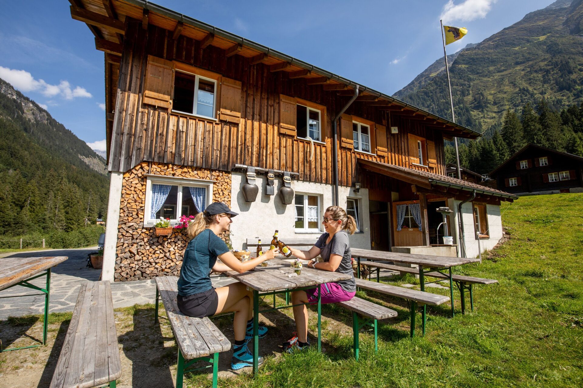 Zwei Personen sitzen an einem Holztisch vor einer rustikalen Berghütte und genießen das sonnige Wetter, während sie Getränke teilen. Die Umgebung vermittelt eine entspannte, freundliche Atmosphäre inmitten einer idyllischen Berglandschaft.