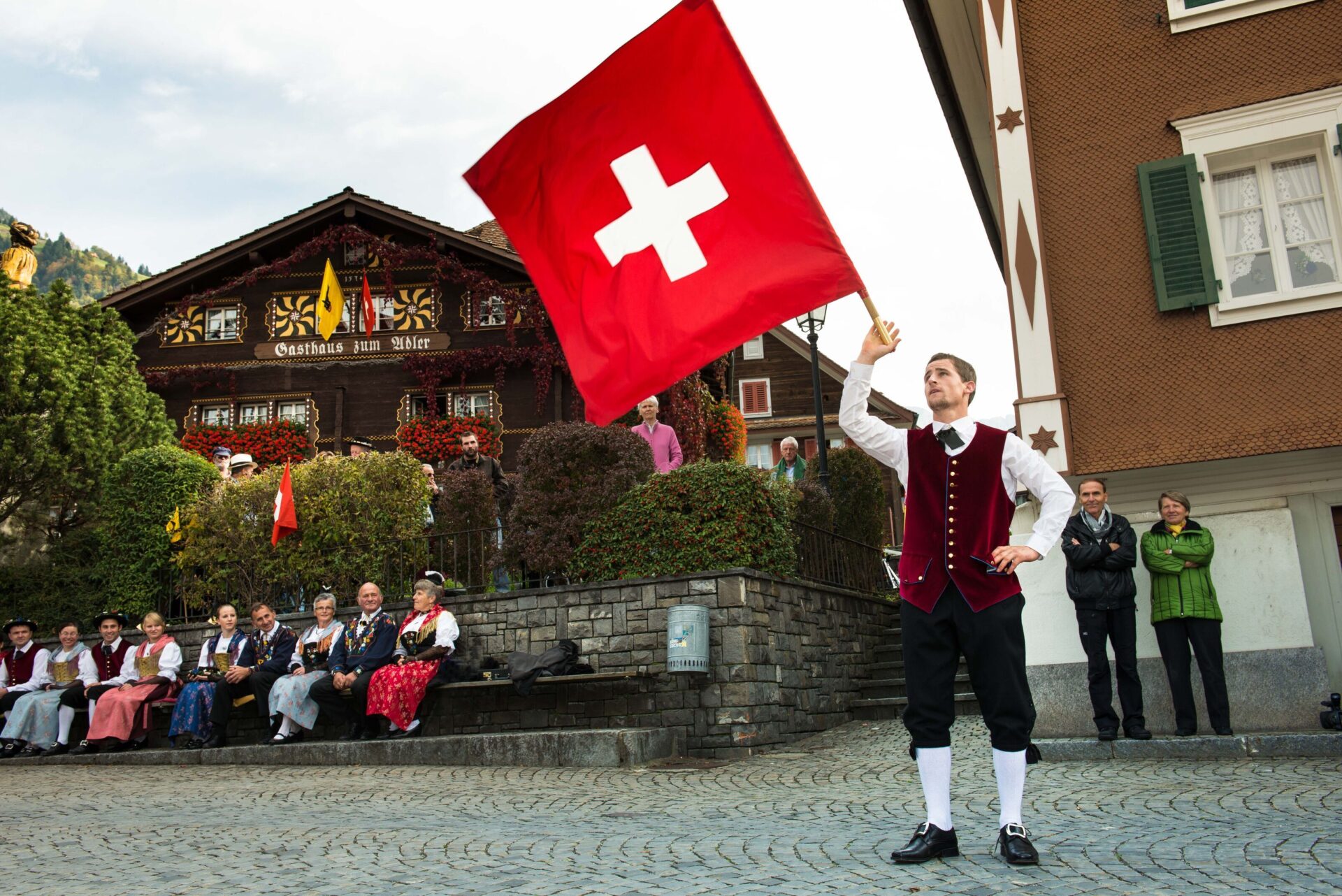 Ein Mann in traditioneller Schweizer Tracht schwenkt eine große Schweizer Fahne auf einem Platz vor einem Chalet, während Zuschauer in Tracht auf einer Steinmauer sitzen und zuschauen.