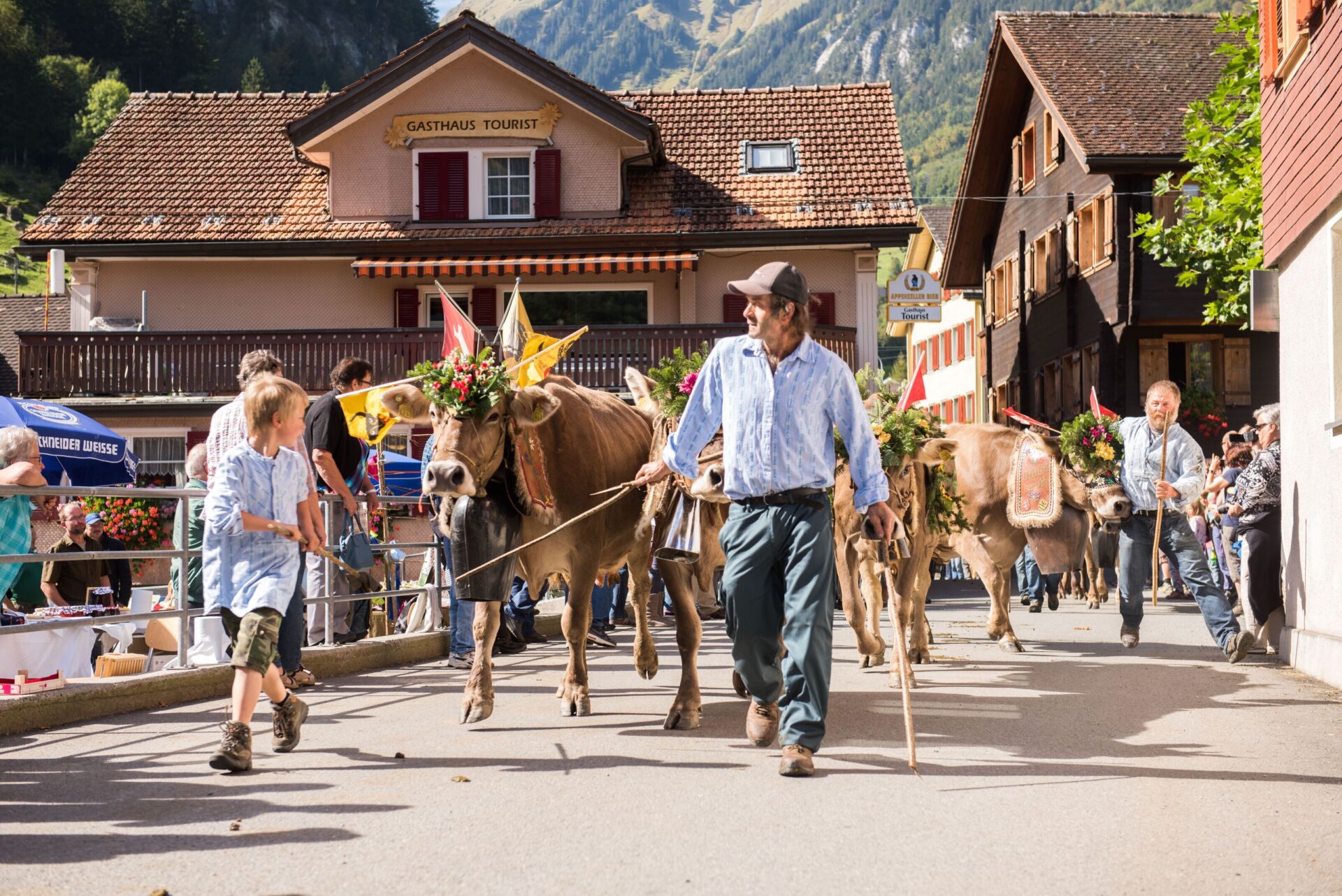 Festlich geschmückte Kühe werden von Bauern und Kindern durch ein Dorf geführt, vermutlich während des traditionellen Alpabzugs. Die Szene strahlt Freude und Gemeinschaft aus, während das ländliche Leben und die Verbundenheit zur alpinen Kultur gefeiert werden.