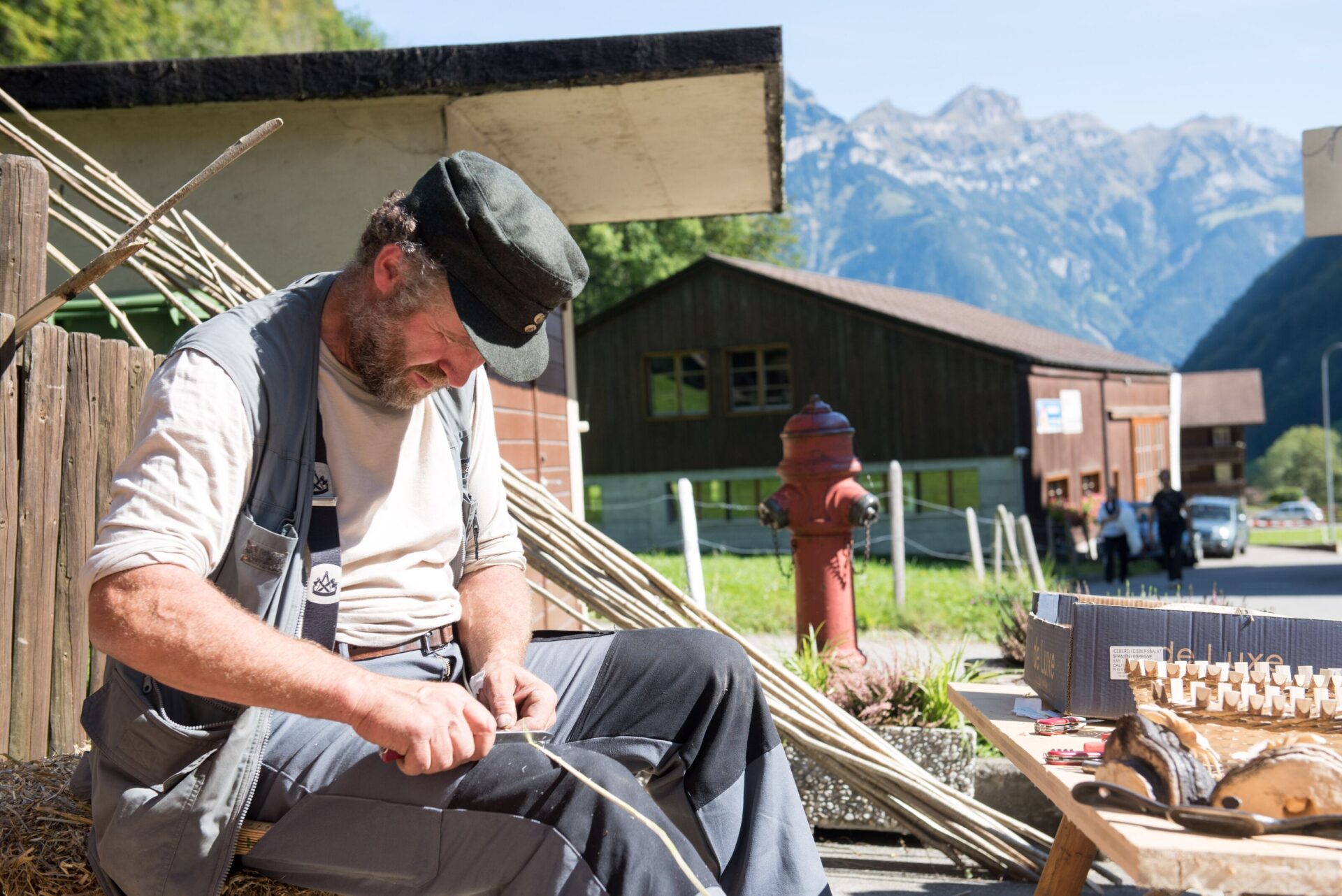 Ein Handwerker sitzt im Freien und bearbeitet konzentriert eine Holzrute, während im Hintergrund eine malerische Berglandschaft und traditionelle Gebäude zu sehen sind. Die Szene vermittelt das Gefühl von handwerklicher Tradition und ländlichem Leben in den Alpen.