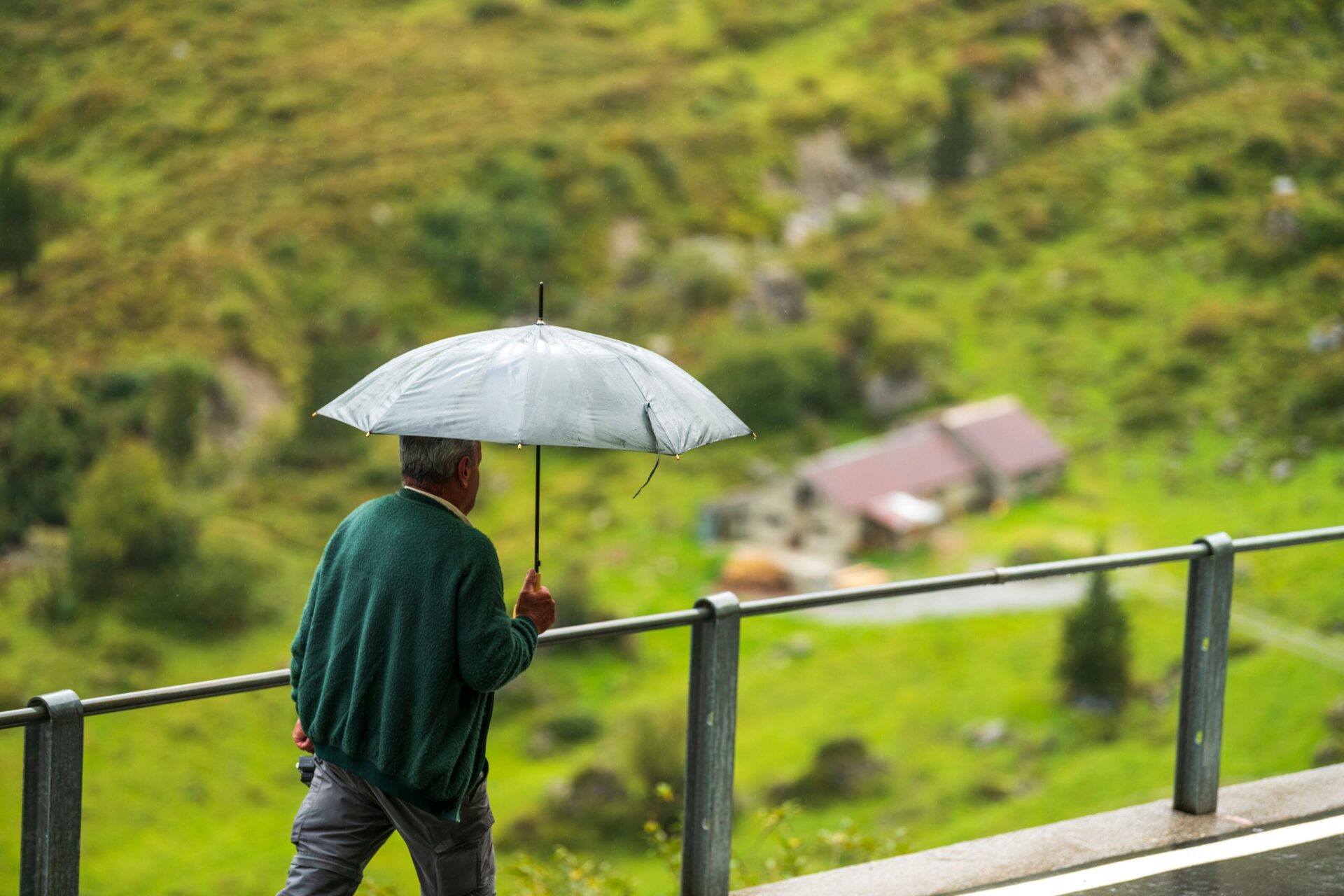 Ein Mann geht mit einem geöffneten Regenschirm entlang eines Geländers, während im Hintergrund eine grüne Berglandschaft mit verstreuten Gebäuden zu sehen ist.