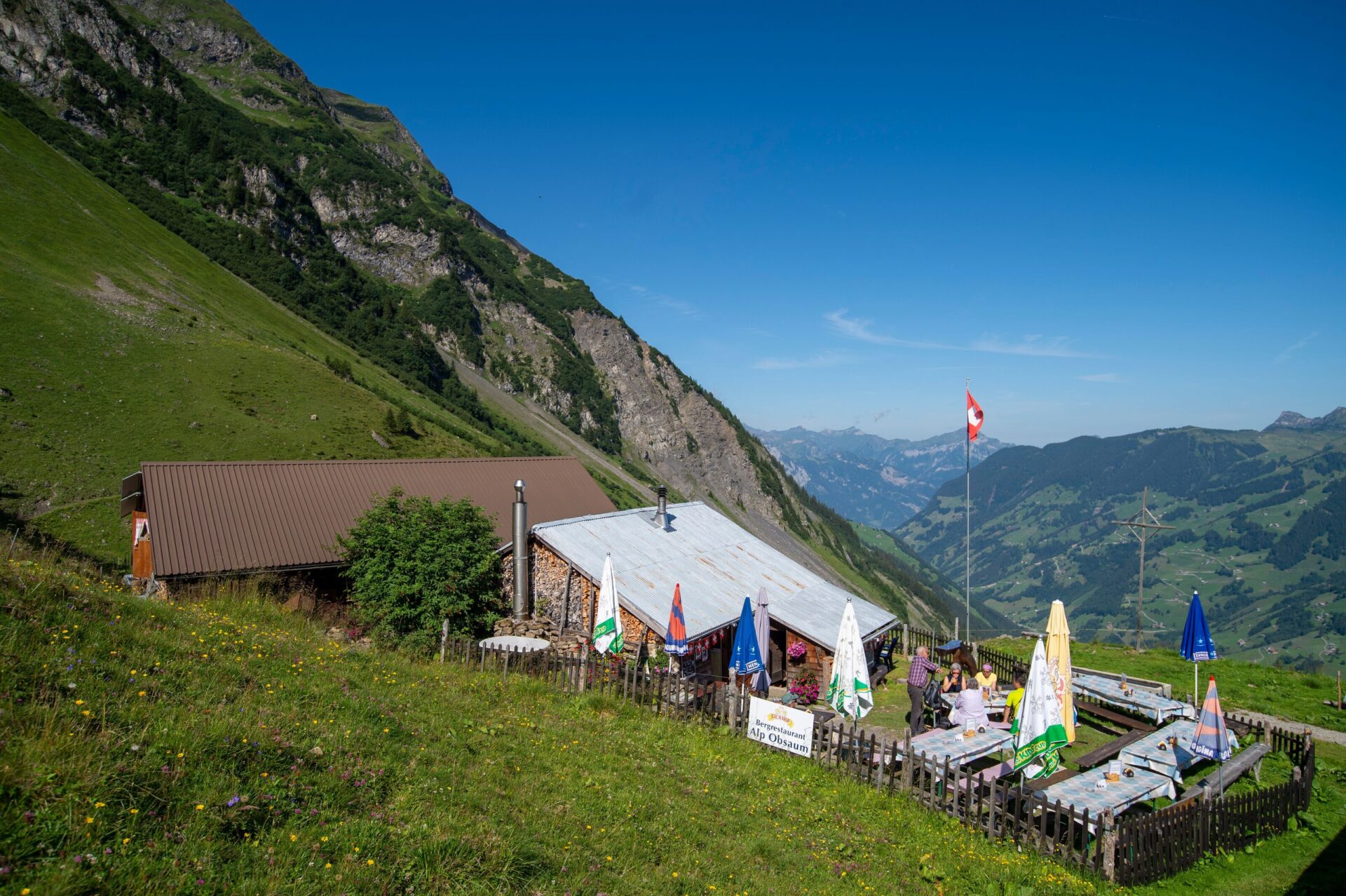 Eine Berghütte mit Sonnenschirmen und Sitzplätzen befindet sich auf einer grünen Wiese, umgeben von hohen Bergen und einem weiten Tal im Hintergrund. Eine Schweizer Fahne weht über der Terrasse.