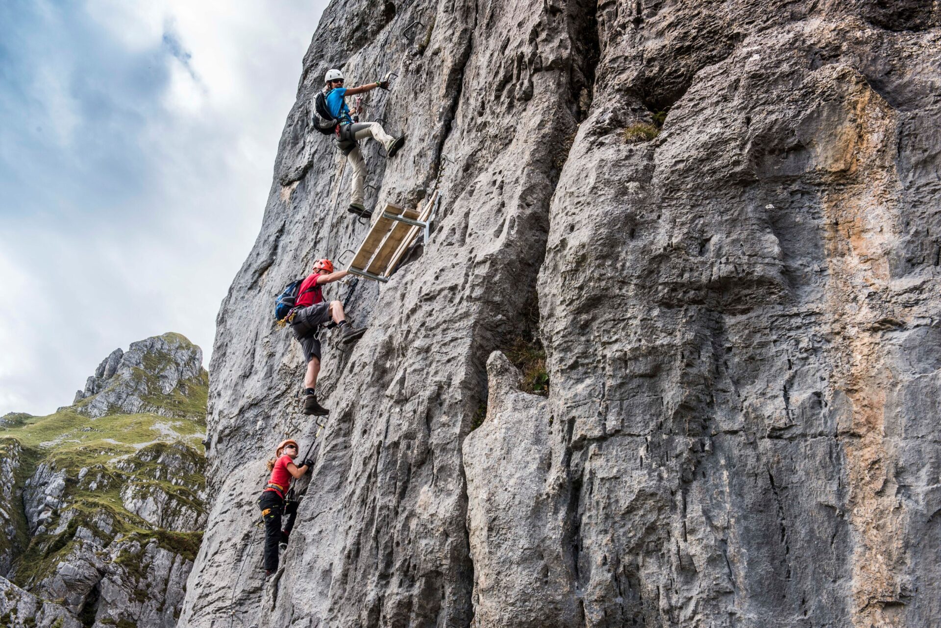 Drei Kletterer mit Sicherheitsausrüstung erklimmen eine steile Felswand, wobei einer eine kleine Plattform passiert. Im Hintergrund sind grüne Berghänge und graue Felsformationen zu sehen, was die alpine Kulisse unterstreicht.