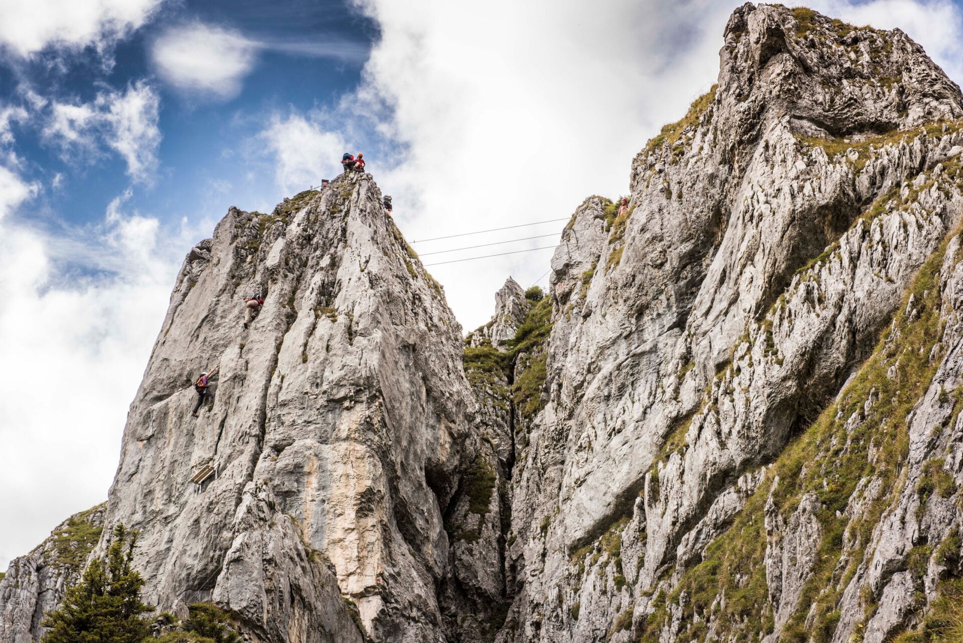 Mehrere Kletterer erklimmen eine steile, zerklüftete Felswand in einer alpinen Landschaft unter einem blauen Himmel mit weißen Wolken. Die Szenerie zeigt die Herausforderung des Kletterns und die atemberaubende Natur der Berge.