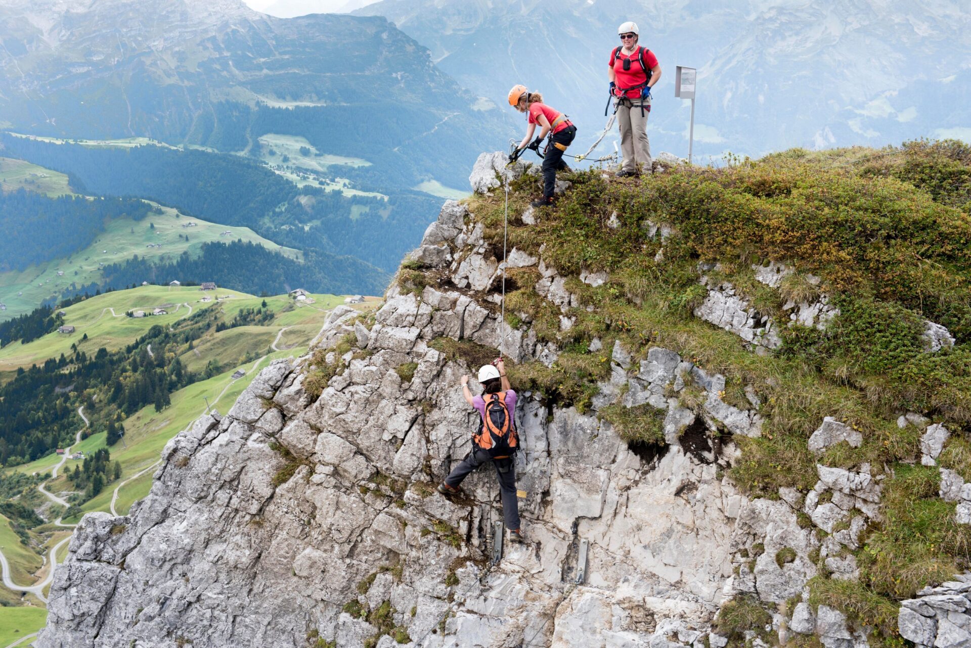Drei Bergsteiger sind an einer felsigen Kante zu sehen, wobei eine Person beim Aufstieg unterstützt wird, während die anderen beiden am Gipfel warten. Die grüne Alplandschaft und verschlungene Wege im Hintergrund schaffen eine beeindruckende, weite Perspektive.