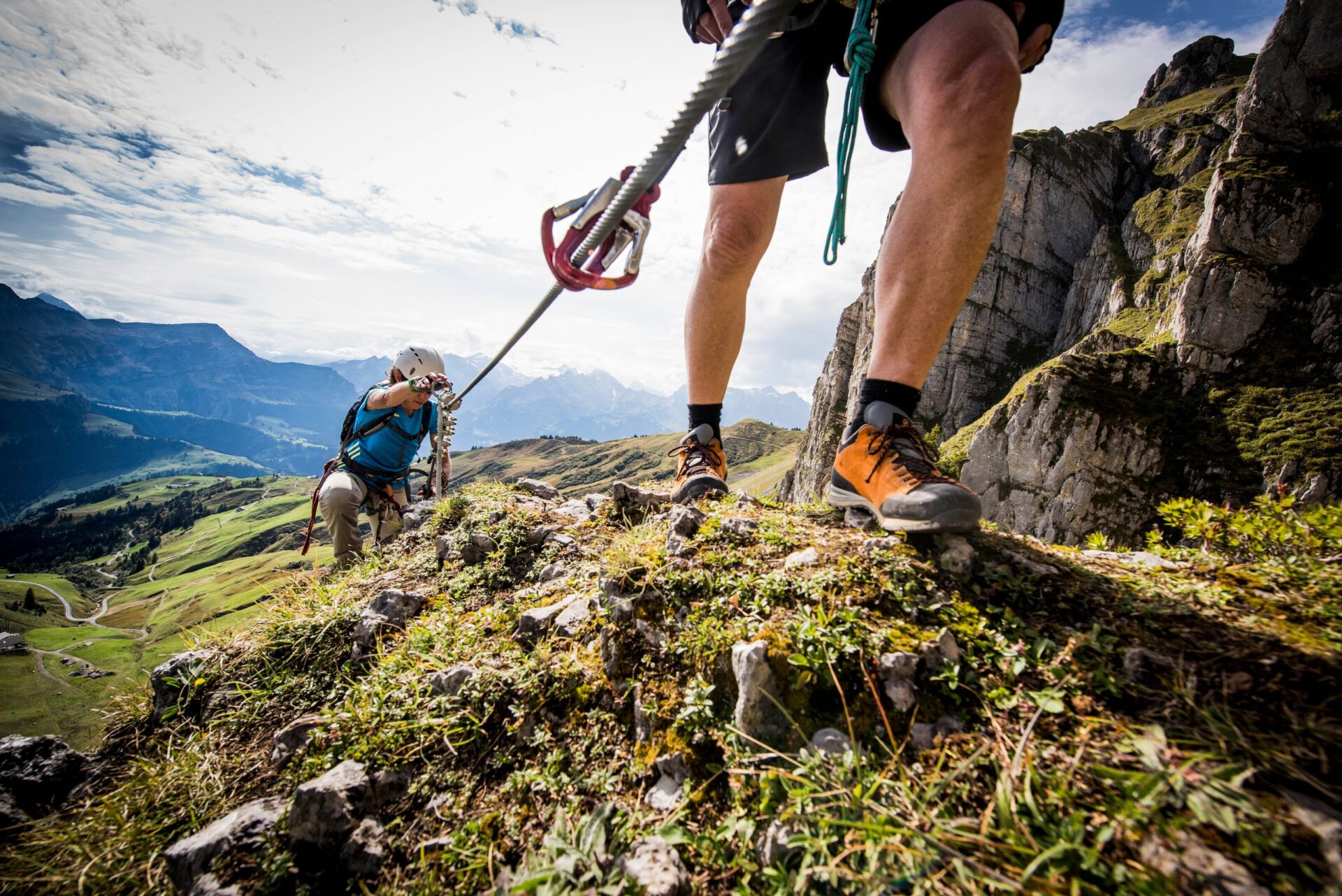 Zwei Bergsteiger erklimmen einen felsigen Klettersteig, gesichert durch ein Stahlseil, während im Hintergrund eine beeindruckende Alpenlandschaft mit grünen Wiesen und schroffen Felsen zu sehen ist. Die Szene strahlt Abenteuer und die Verbundenheit mit der Natur aus, unterstrichen durch den sonnigen Himmel und die klare Bergluft.