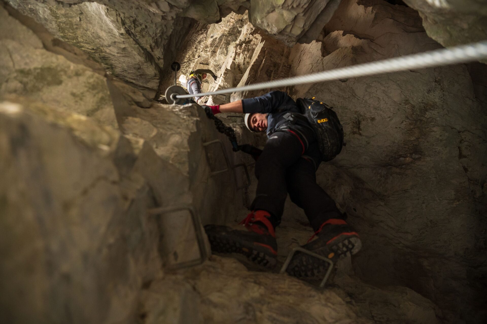 Ein Kletterer mit Helm und Rucksack erklimmt eine steile, felsige Wand in einer Höhle, gesichert durch ein Stahlseil und Metalltritte. Das Bild vermittelt Spannung und die Herausforderung eines anspruchsvollen Klettersteigs.