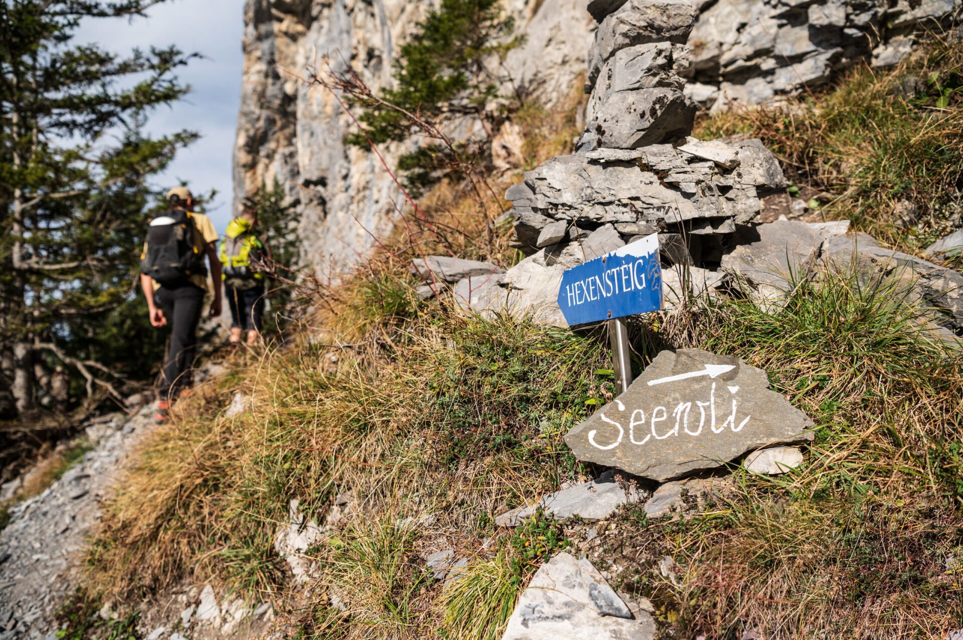 Ein Wegweiser mit den Aufschriften „Hexensteig“ und „Seewli“ steht inmitten einer felsigen und grasbewachsenen Berglandschaft, während zwei Wanderer mit Rucksäcken dem Pfad folgen. Die Szene strahlt Abenteuerlust und Naturverbundenheit aus.