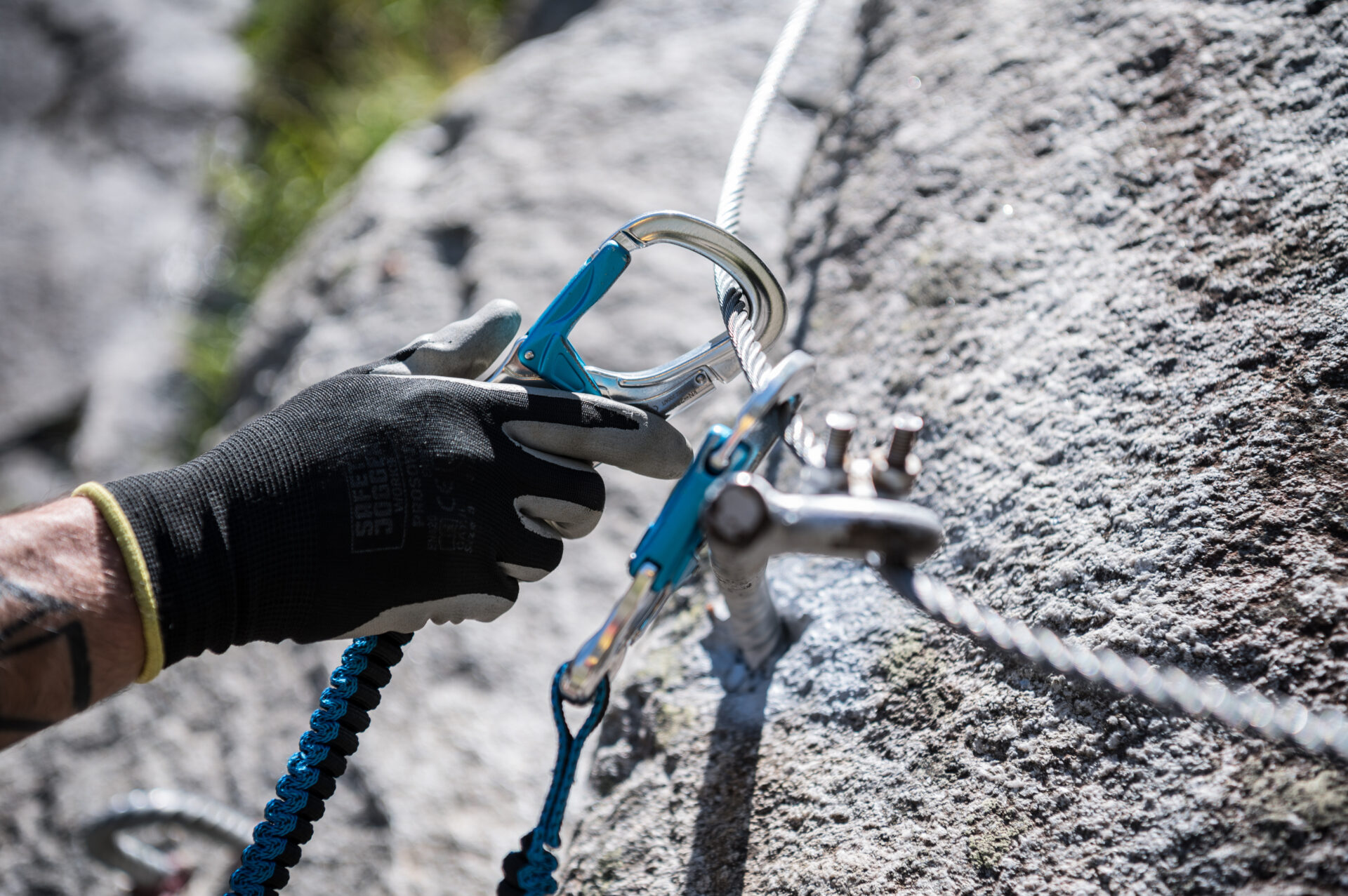 Eine behandschuhte Hand sichert einen blauen Karabiner an einem Stahlseil, das an einer felsigen Oberfläche befestigt ist, während die Sicherheitsausrüstung für Klettersteige im Fokus steht. Das Bild vermittelt Präzision und Sicherheit beim Klettern in steilem Gelände.