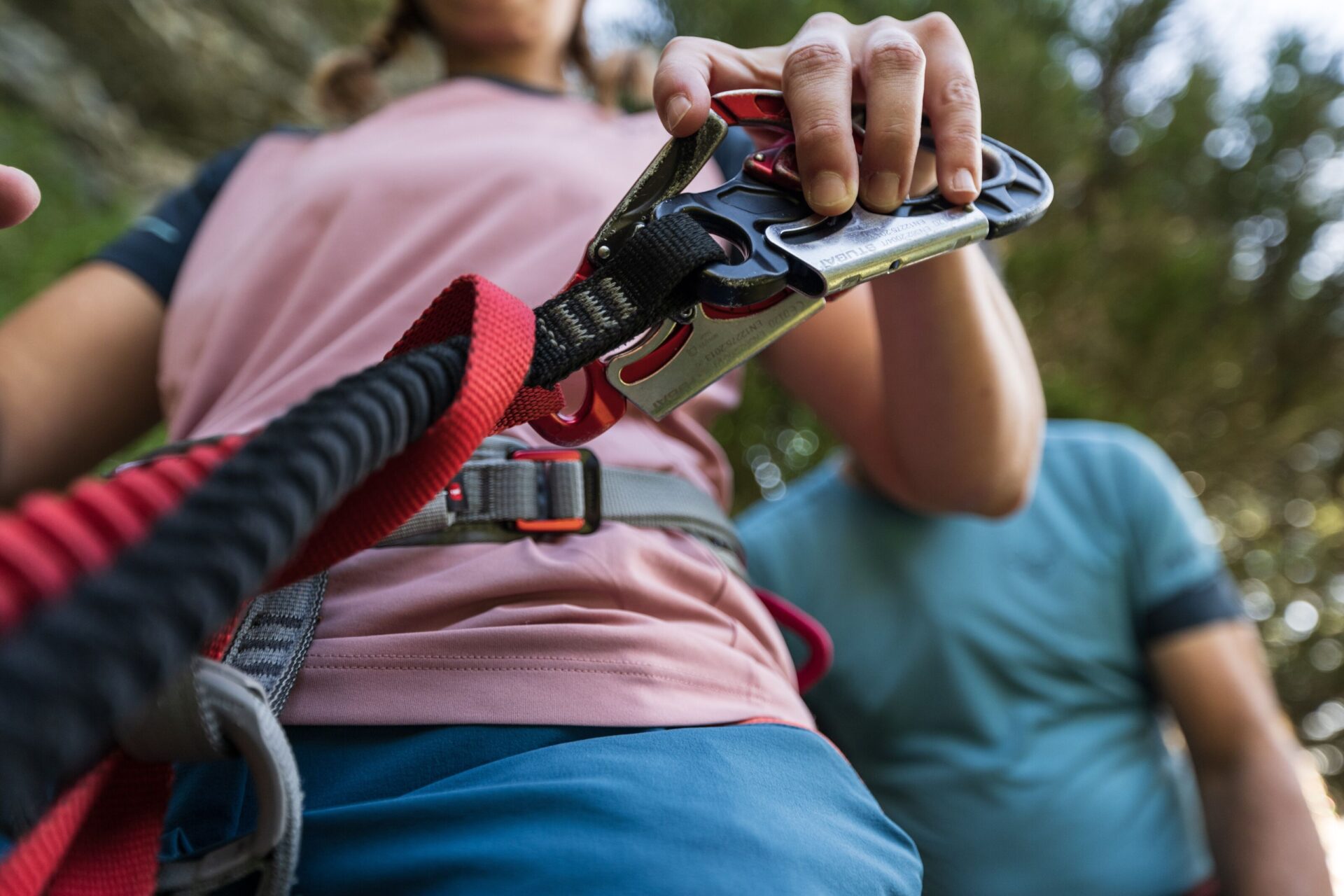 Das Bild zeigt eine Nahaufnahme einer Person, die einen Kletterkarabiner an einem Sicherheitsseil befestigt, mit Fokus auf die Hände und die Ausrüstung. Die Umgebung deutet auf eine Outdoor-Kletterszene hin, mit unscharfen Bäumen im Hintergrund.