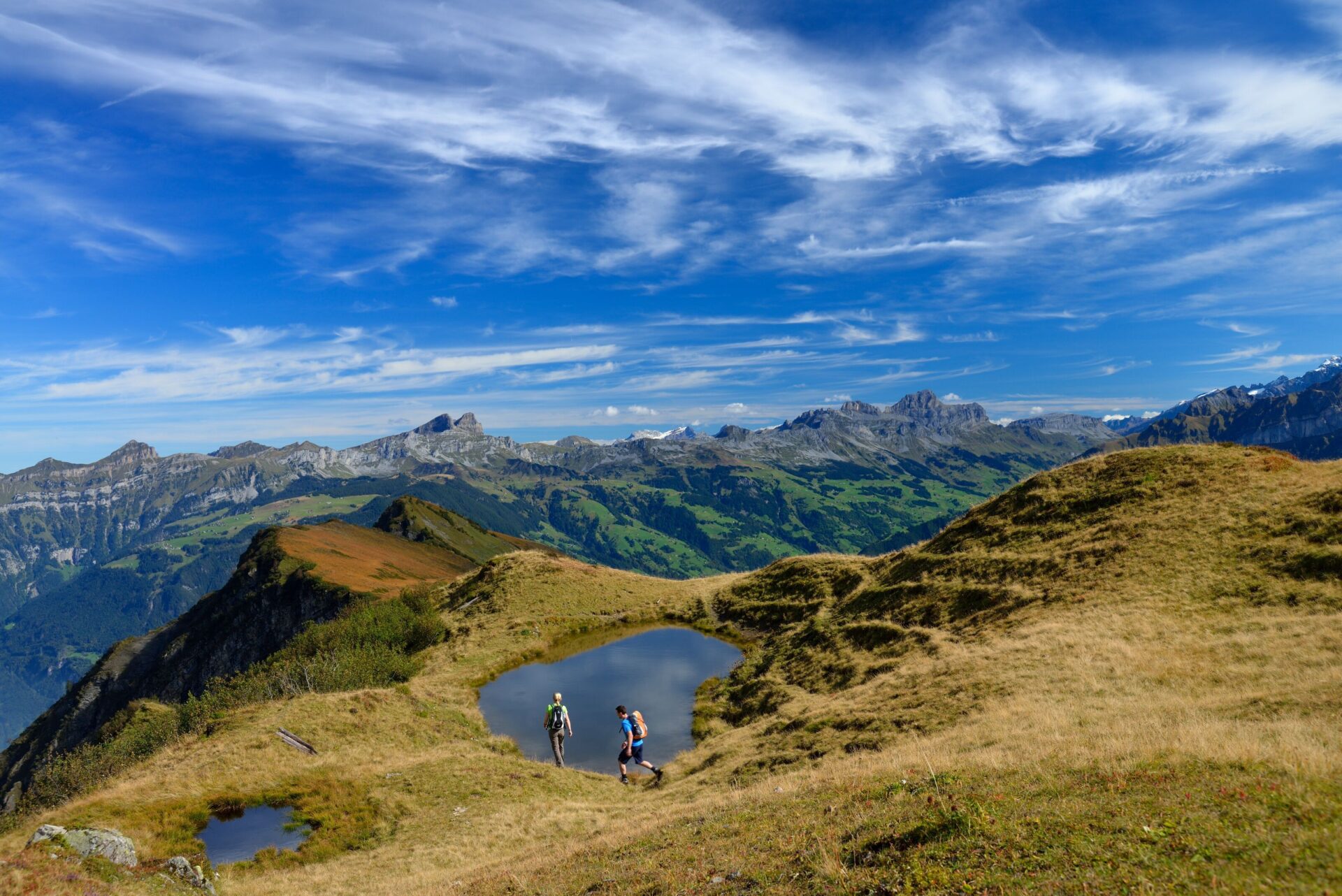 Zwei Wanderer stehen an einem kleinen Bergsee auf einem grasbewachsenen Hochplateau, umgeben von einer spektakulären Alpenkulisse unter einem klaren, blau-weißen Himmel. Die Szene strahlt Weite, Freiheit und die Schönheit der Bergnatur aus.