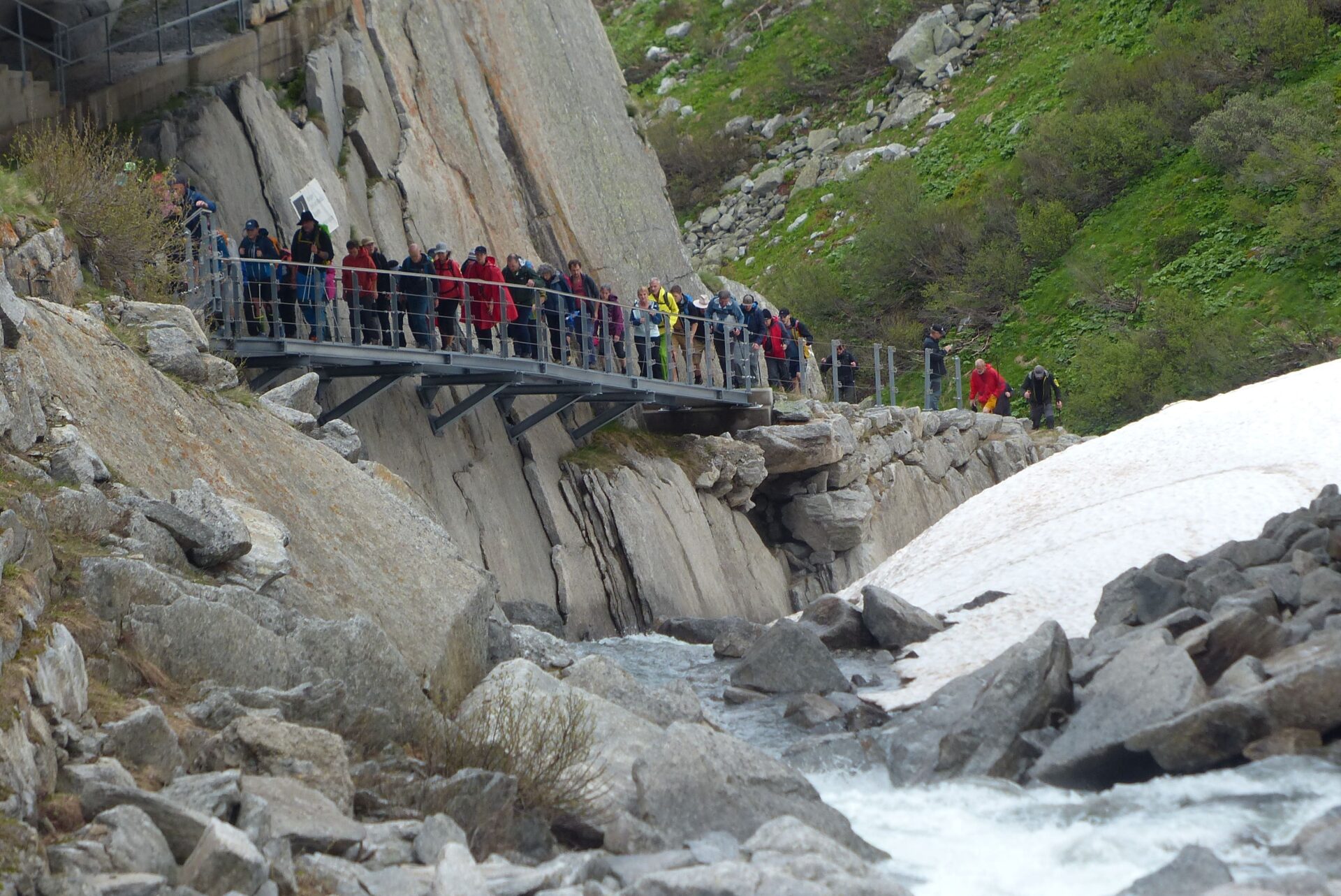 Eine Gruppe von Wanderern überquert einen schmalen Steg entlang steiler Felswände, während unter ihnen ein Gebirgsbach rauscht. Die wilde Berglandschaft und der Schnee betonen die Abenteuerlust auf dem Gottardo-Wanderweg.