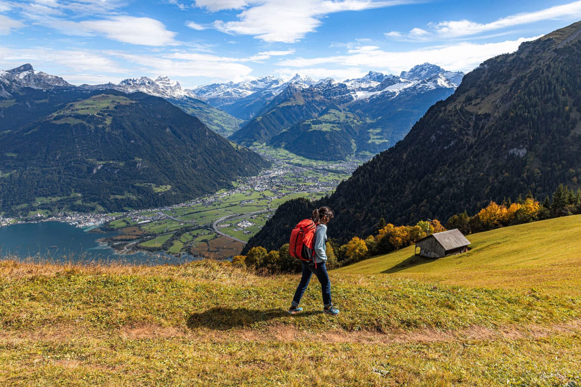 Ein Wanderer mit rotem Rucksack läuft einen grasigen Pfad entlang, mit einem atemberaubenden Blick auf ein Tal, einen See und die umliegenden Berge. Die herbstlich gefärbte Landschaft und der weite Himmel verstärken das Gefühl von Freiheit und Naturverbundenheit.