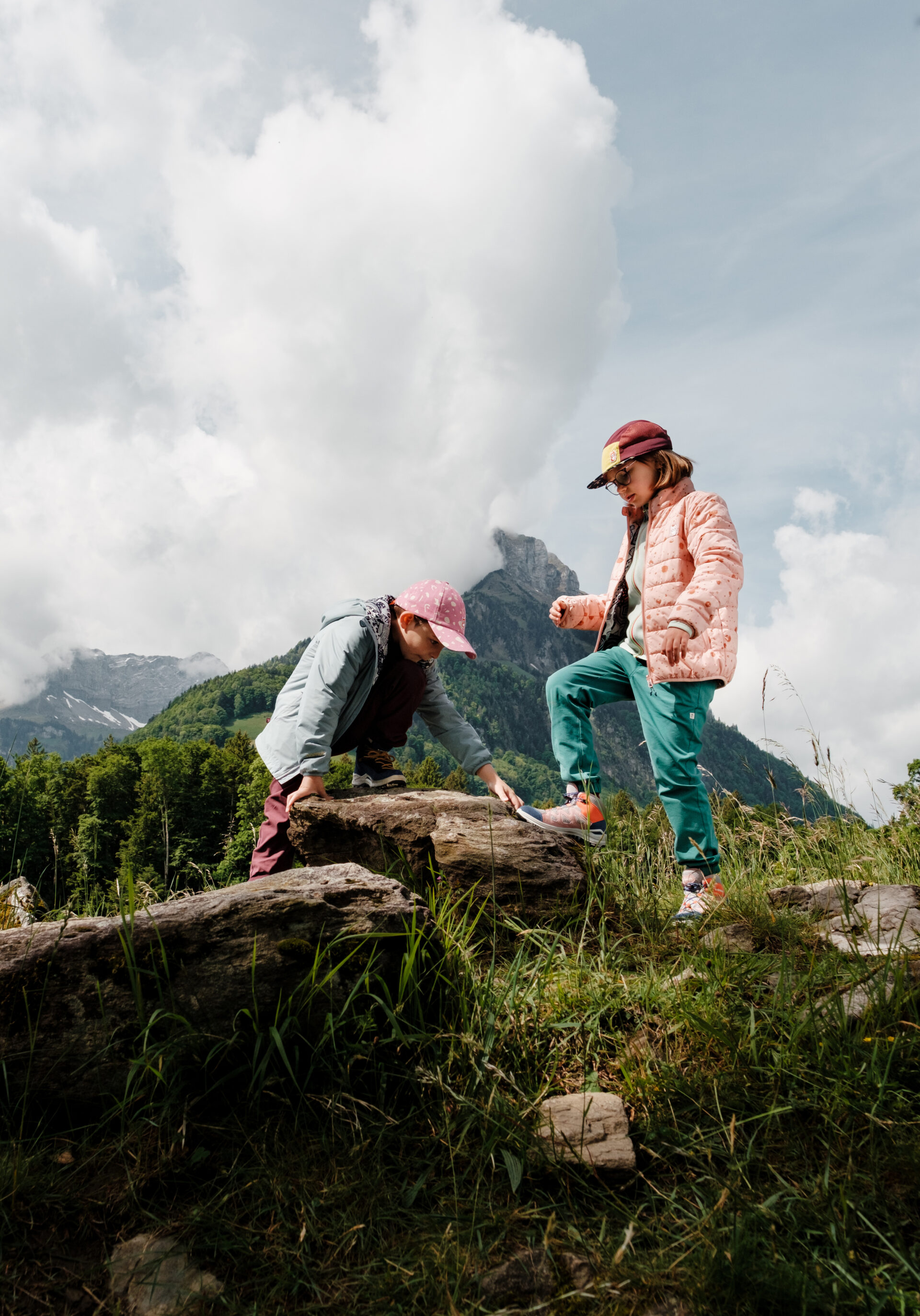 Zwei Kinder in farbenfroher Outdoor-Kleidung erkunden spielerisch eine Wiese mit Felsen in einer alpinen Berglandschaft. Im Hintergrund ragen grüne Hänge und ein markanter Gipfel unter großen, hellen Wolken in den Himmel und verleihen der Szene eine lebendige, abenteuerliche Stimmung.