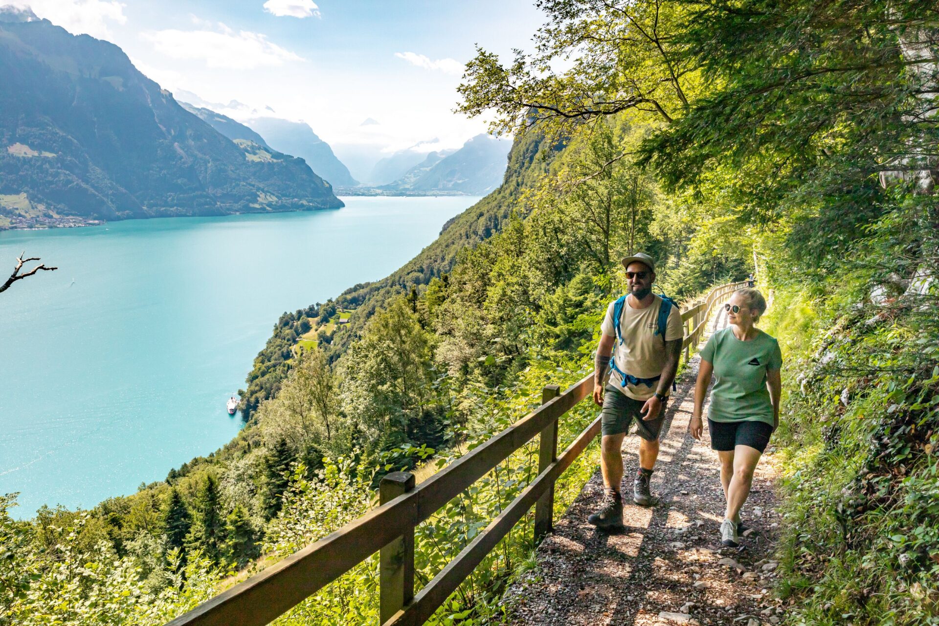 Zwei Wanderer spazieren auf einem schmalen Pfad entlang eines Hügels mit Blick auf einen türkisblauen See und die umgebenden Berge. Die sonnige Landschaft und der ruhige Weg vermitteln ein Gefühl von Entspannung und Naturverbundenheit.